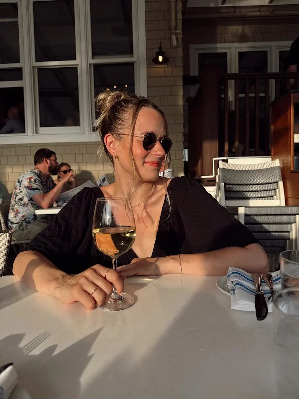 A woman with sunglasses and a bun hairstyle, sitting at an outdoor table, holding a glass of white wine, smiling, with people in the background, some wearing patterned shirts, in a casual dining setting during daytime.