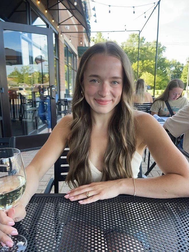A young woman with long wavy brown hair sitting at an outdoor cafe table with a glass of white wine, smiling at the camera. Other diners and green trees are visible in the background.