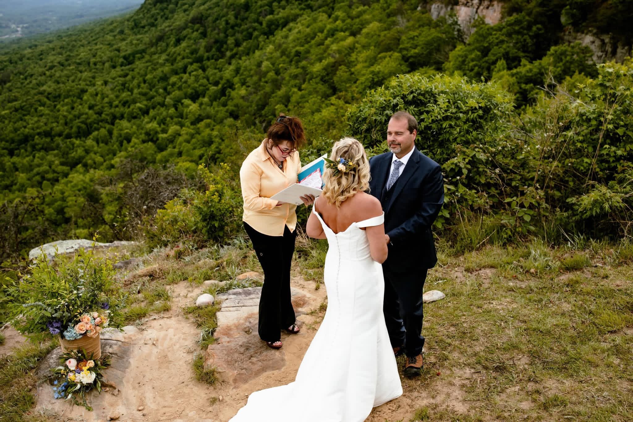 A wedding ceremony outdoors on a hillside with greenery and mountains in the background, featuring a bride, a groom, and an officiant.