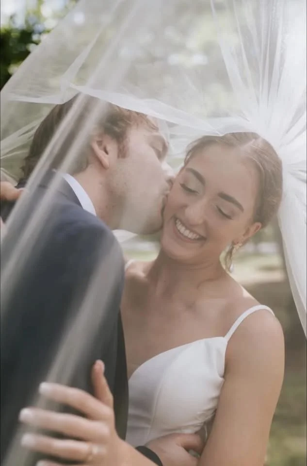 A newlywed couple sharing a kiss under a clear umbrella outdoors, smiling and embracing each other.
