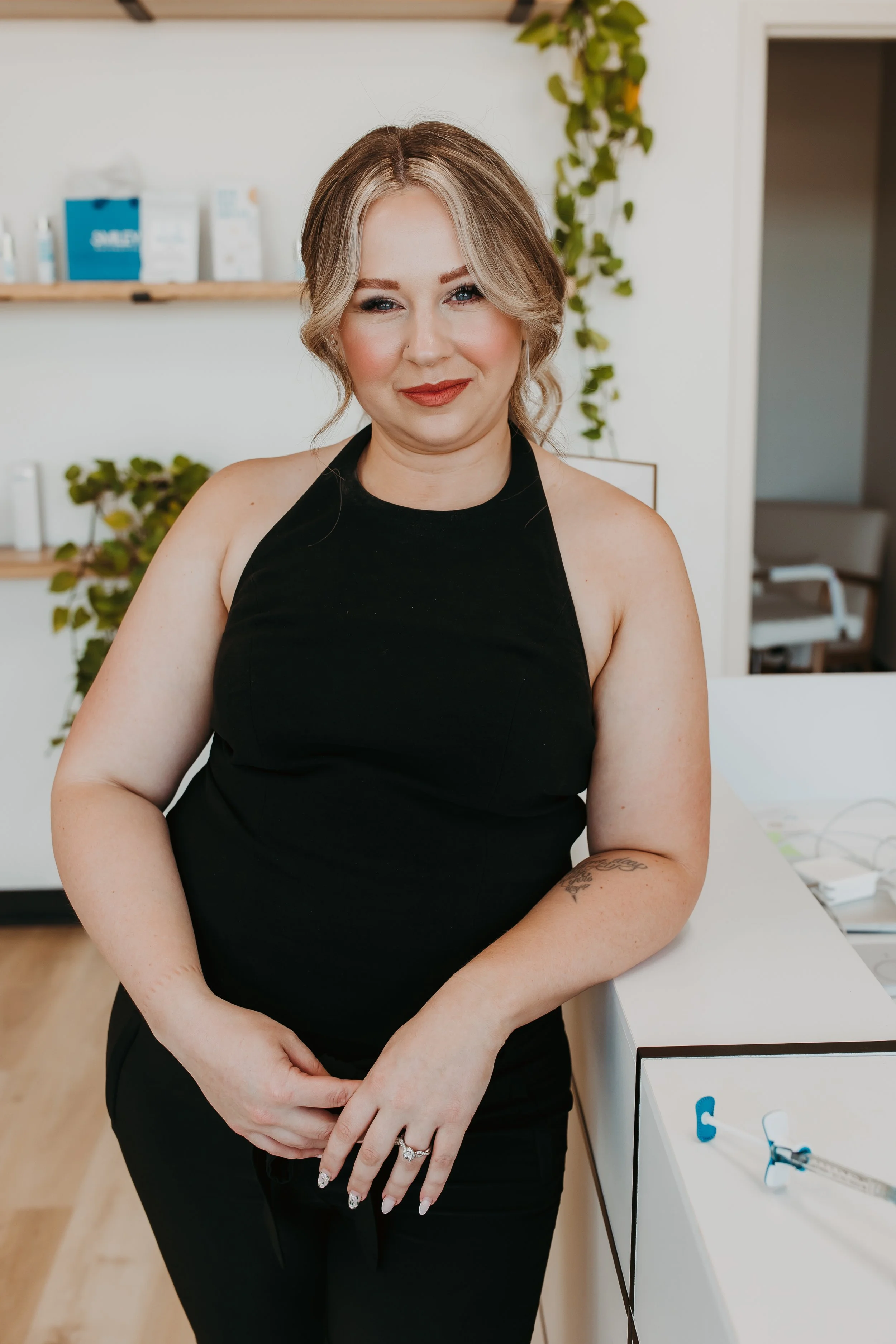 A woman with blonde hair styled in loose waves, wearing a sleeveless black dress, standing in a modern interior space with white walls and wooden floors. She is smiling gently, showing her red lipstick and subtle eye makeup, with her hands clasped in front of her. There are green plants and shelves with products in the background.