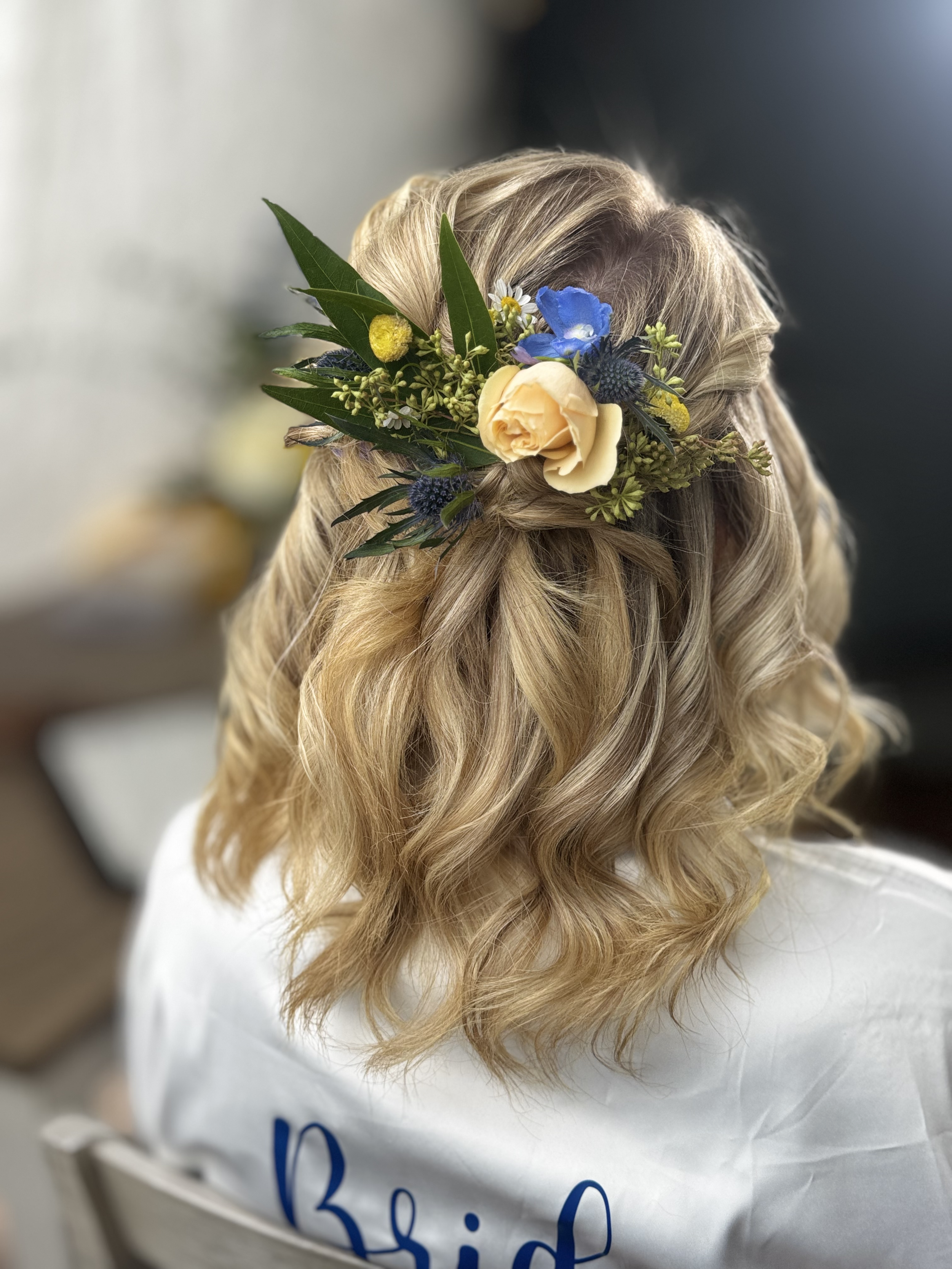 A person's blonde hair styled in loose curls with a floral hair accessory, including a peach rose, blue flowers, and green leaves.