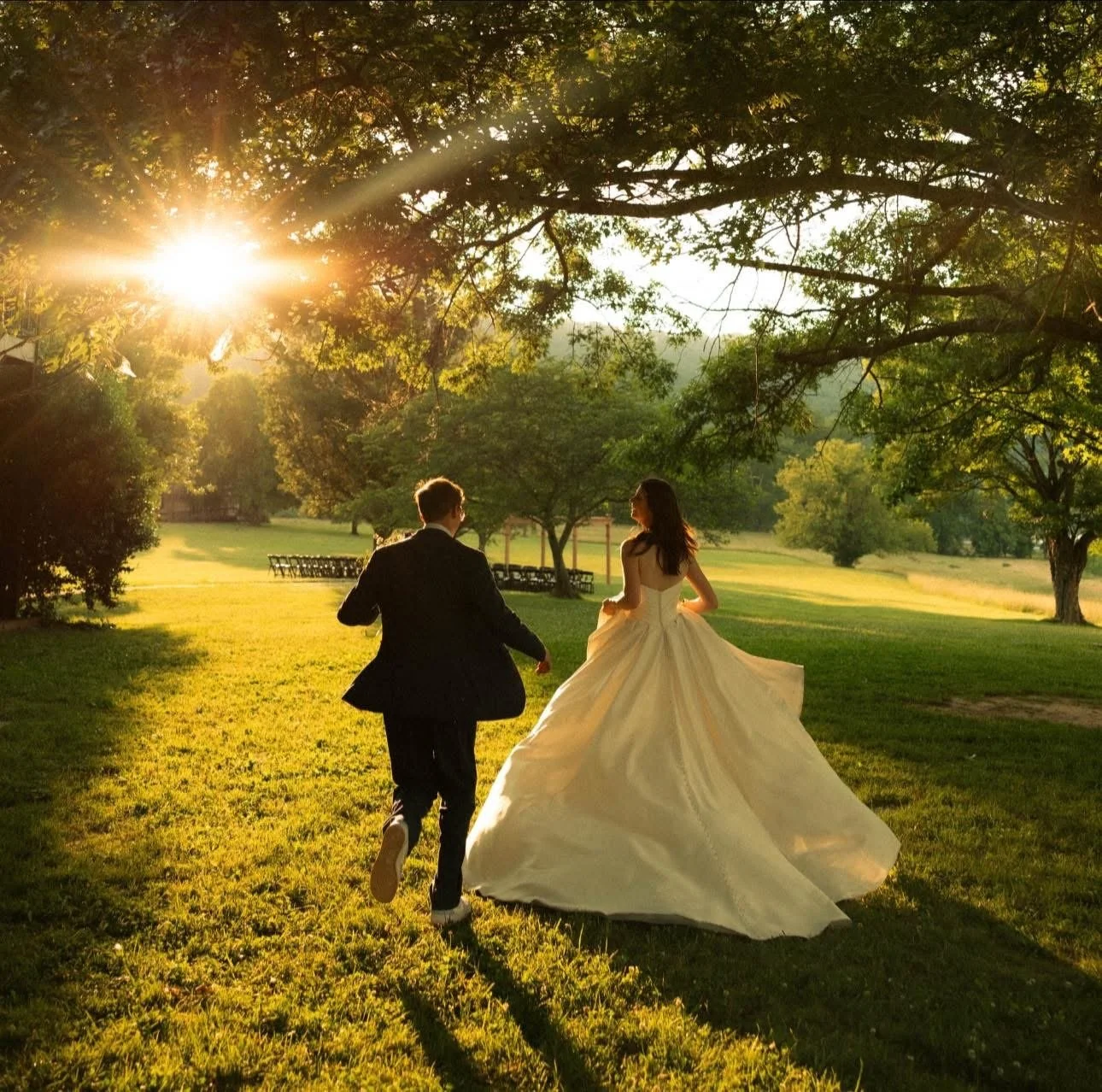 A bride in a white wedding gown and a groom in a black suit run together in a park at sunset, with trees and green grass surrounding them.