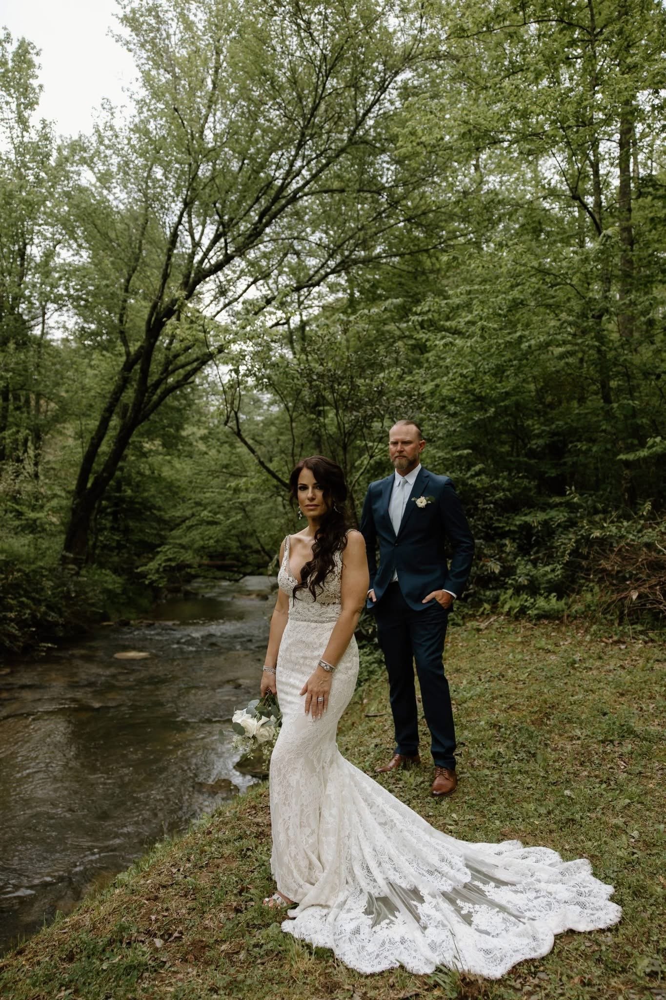 A bride in a white lace wedding gown with a long train holding a bouquet of white flowers and greenery, standing barefoot on grass near a small stream, with a groom in a dark blue suit and brown shoes standing behind her in a lush green forest.