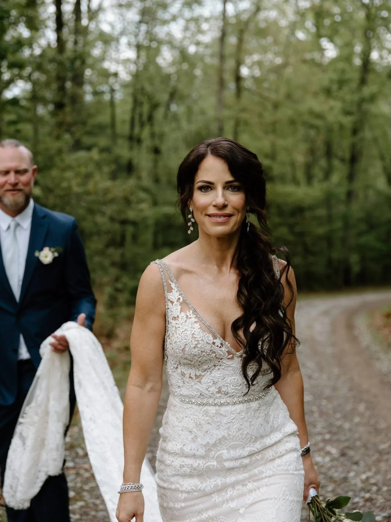 A woman in a lace wedding dress walking outdoors on a wooded path, smiling, with a man in a suit walking behind her holding a white garment.