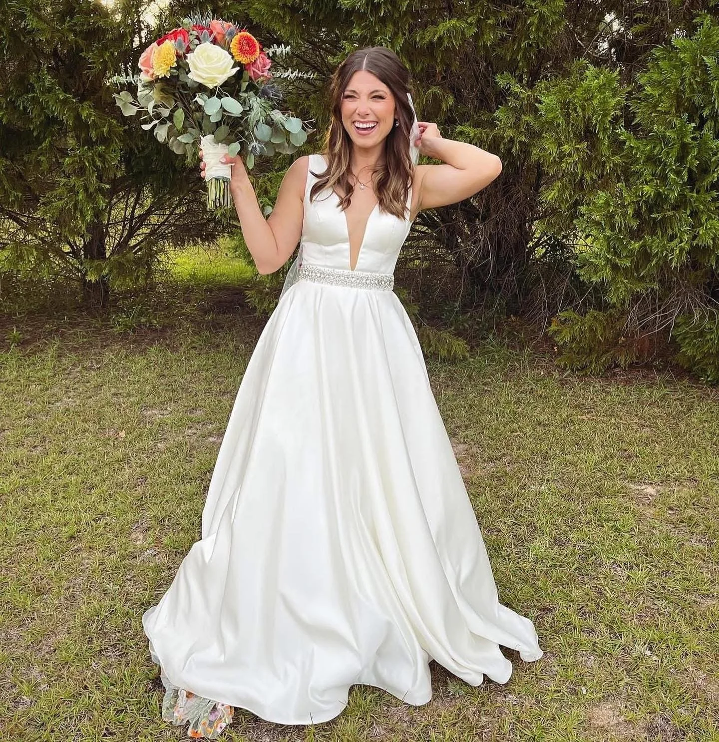 A woman in a white wedding dress holding a large bouquet of flowers, standing on grass in front of green bushes, smiling and laughing.