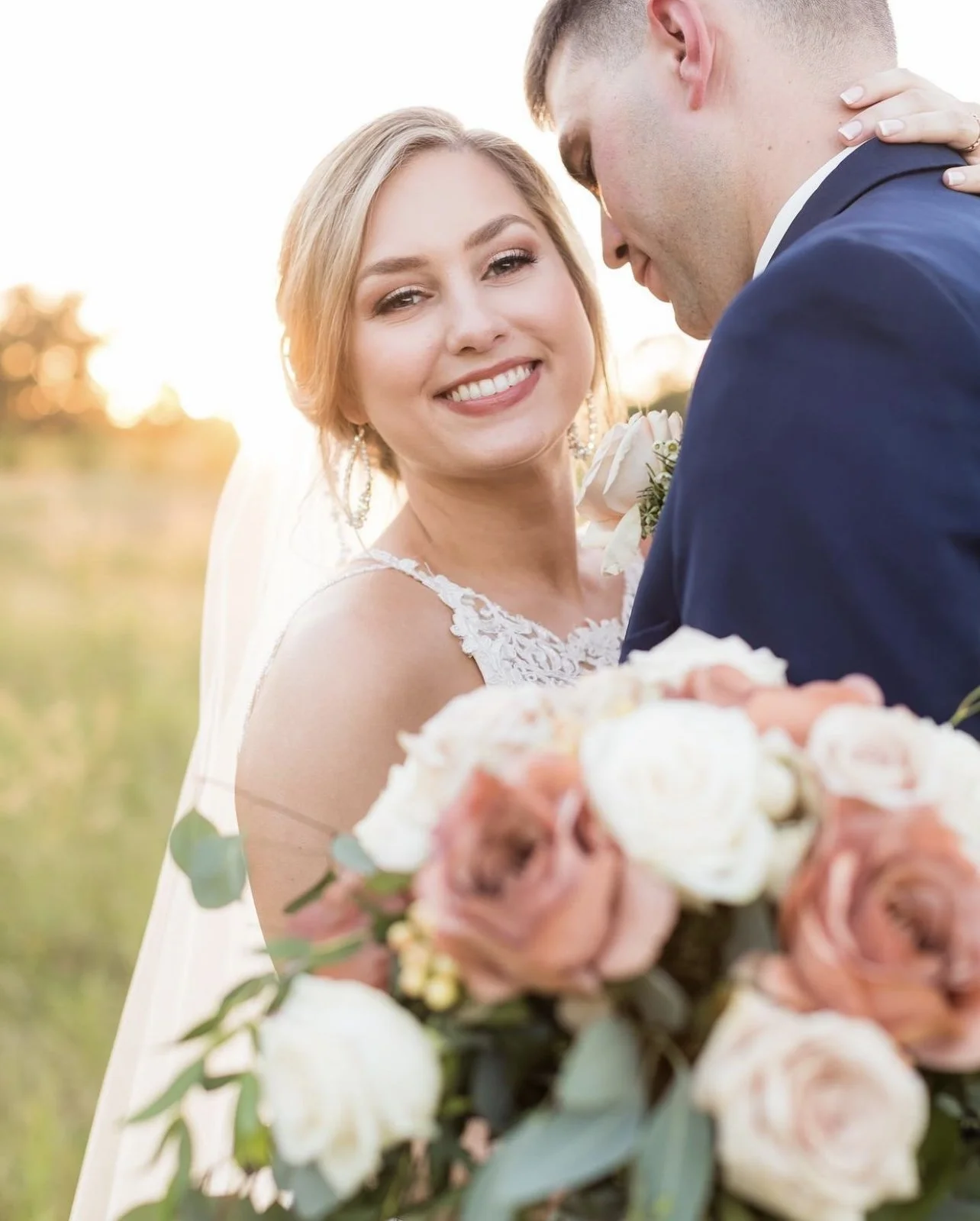 A smiling bride and groom on their wedding day outdoors during sunset, with the bride holding a bouquet of pink and white roses and the groom leaning in for a kiss.