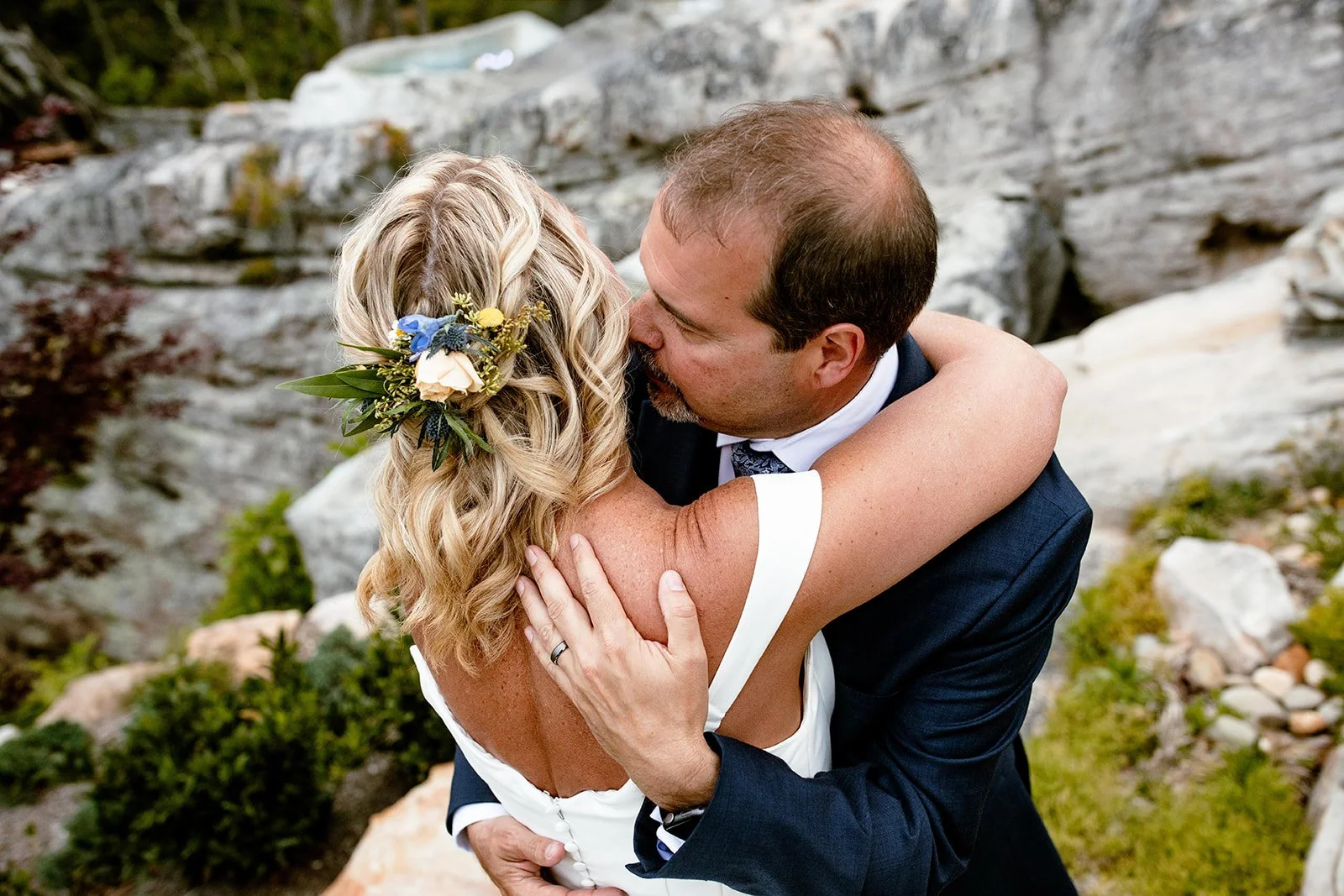 A couple hugging and kissing outdoors, the woman with blonde hair and a floral hairpiece, and the man in a dark suit and tie, surrounded by rocks and greenery.