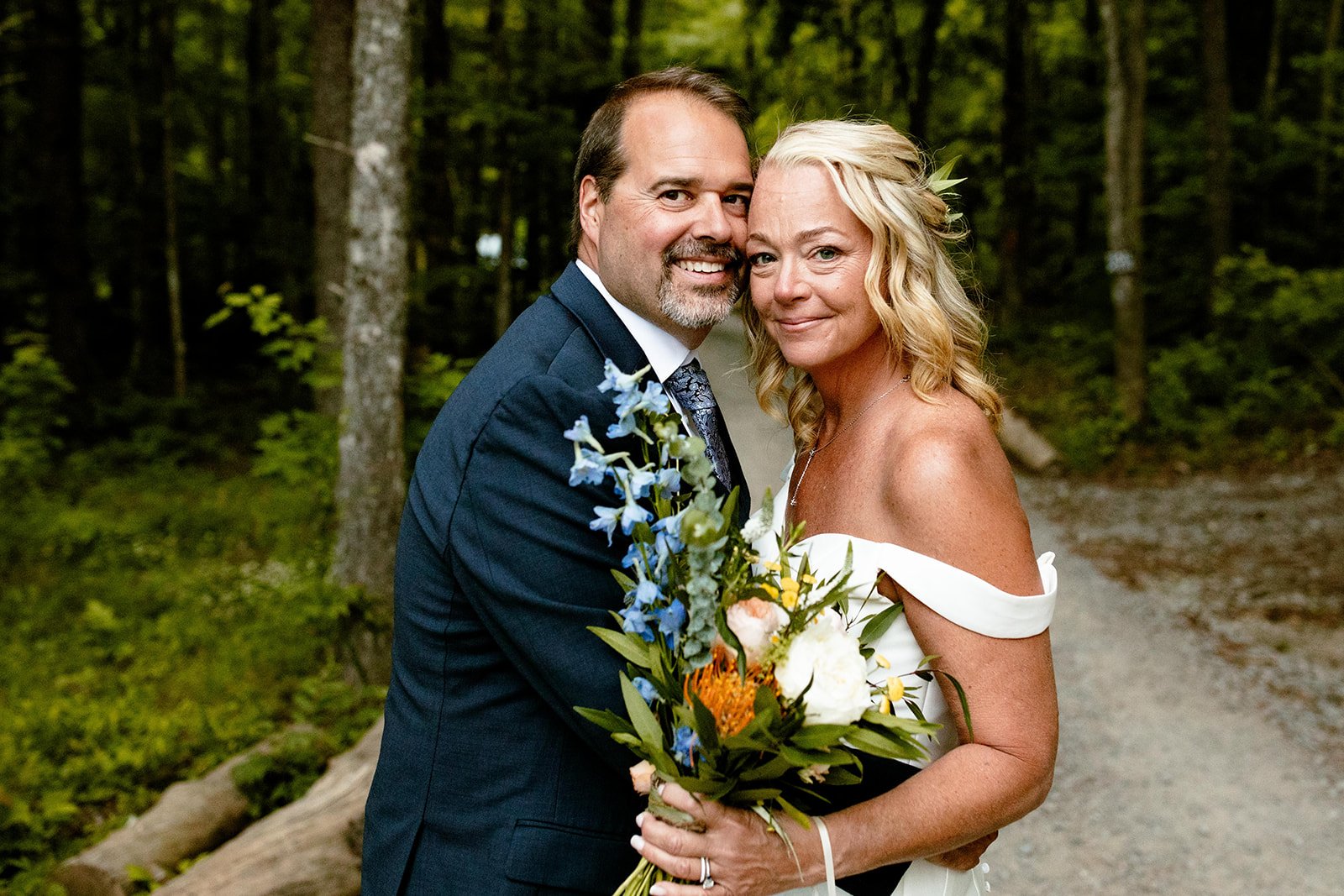 A couple dressed in wedding attire embracing outdoors in a wooded area, holding a bouquet of flowers.