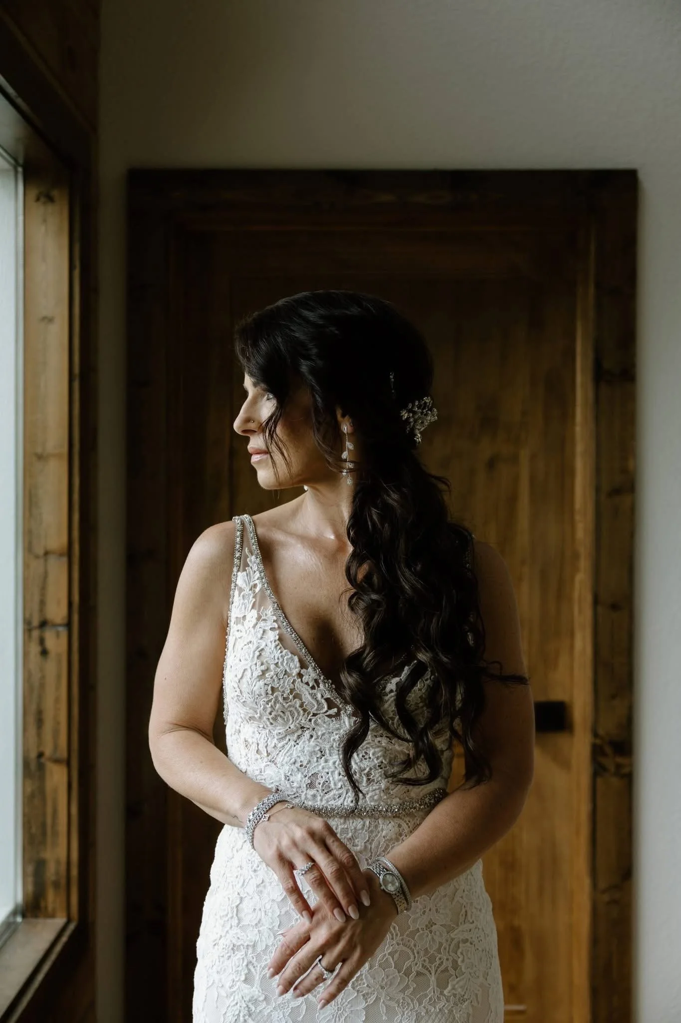 A woman with long dark hair styled in loose waves, dressed in a lace wedding gown with a V-neckline, standing near a window with natural light. She is wearing elegant jewelry, including earrings, a bracelet, and a ring, and is looking to her left wit