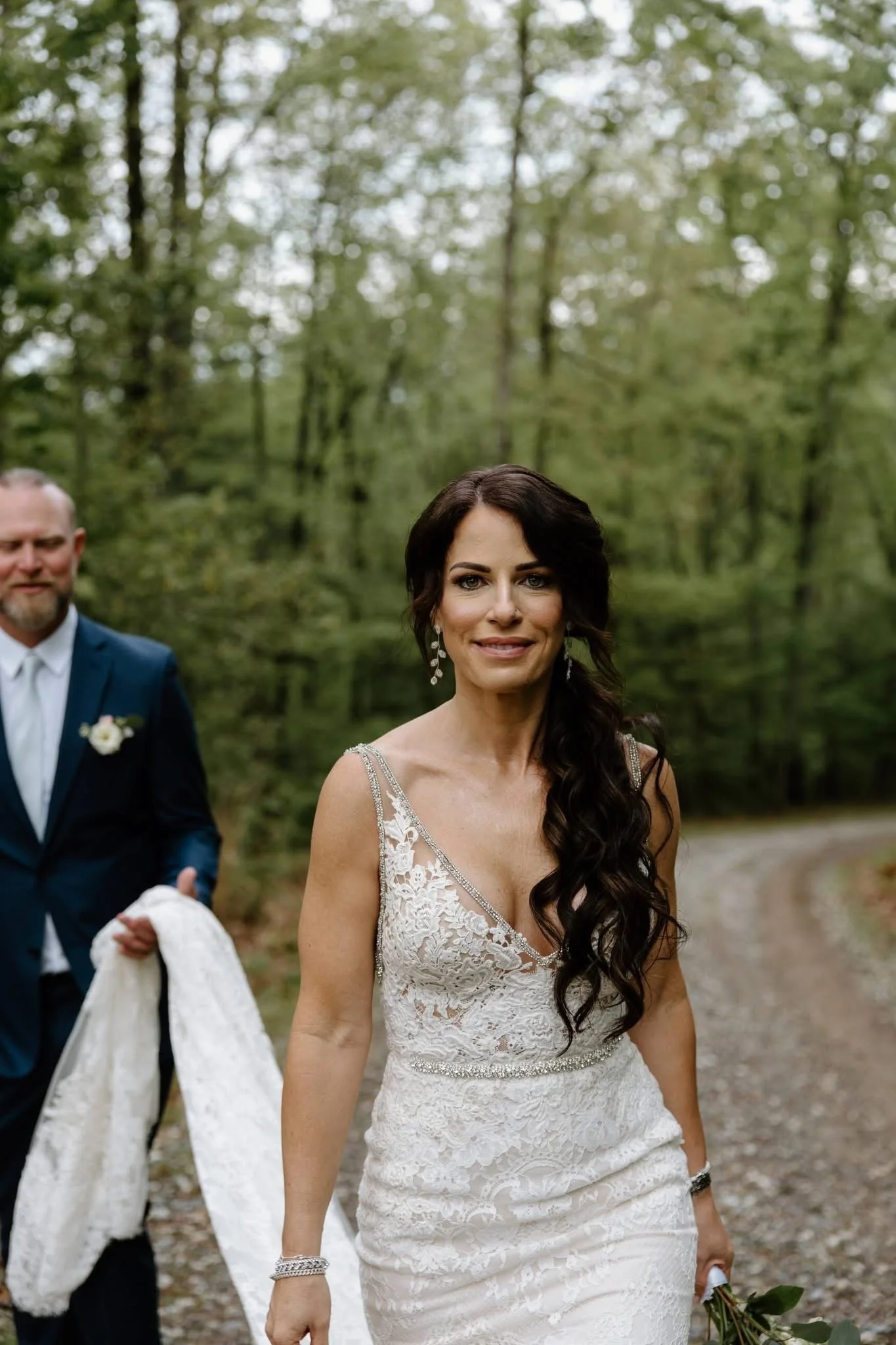 A bride with long dark hair in a white lace wedding dress smiling outdoors, walking with a groom in a blue suit.
