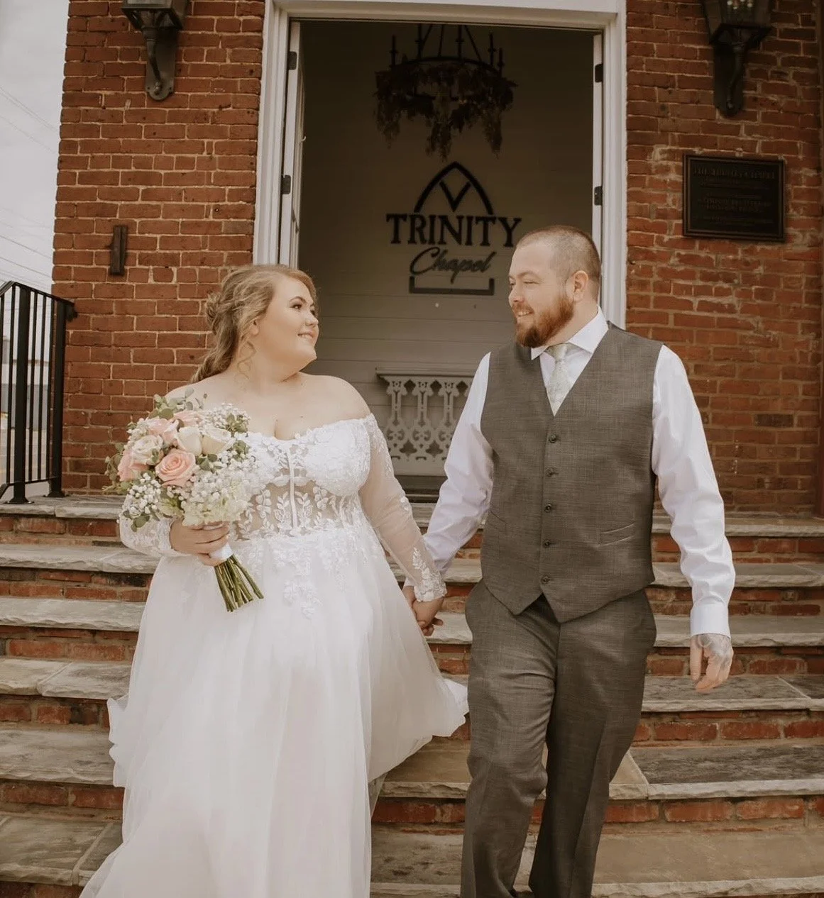 A bride and groom holding hands and smiling at each other outside Trinity Chapel, with brick steps and a chandelier inside visible in the background.