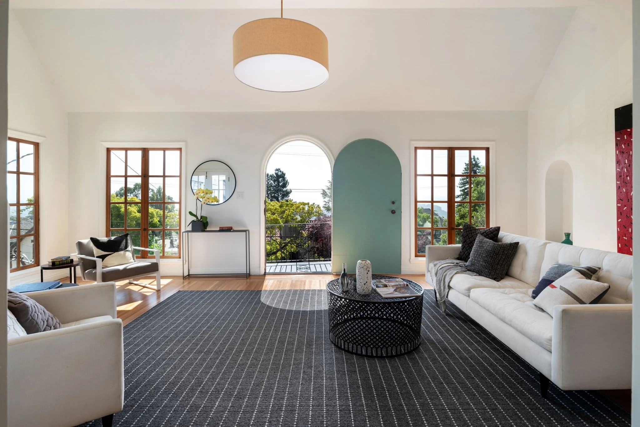 Bright living room with large windows, white sofas, a black coffee table, and a view of trees outside through an arched door and windows.