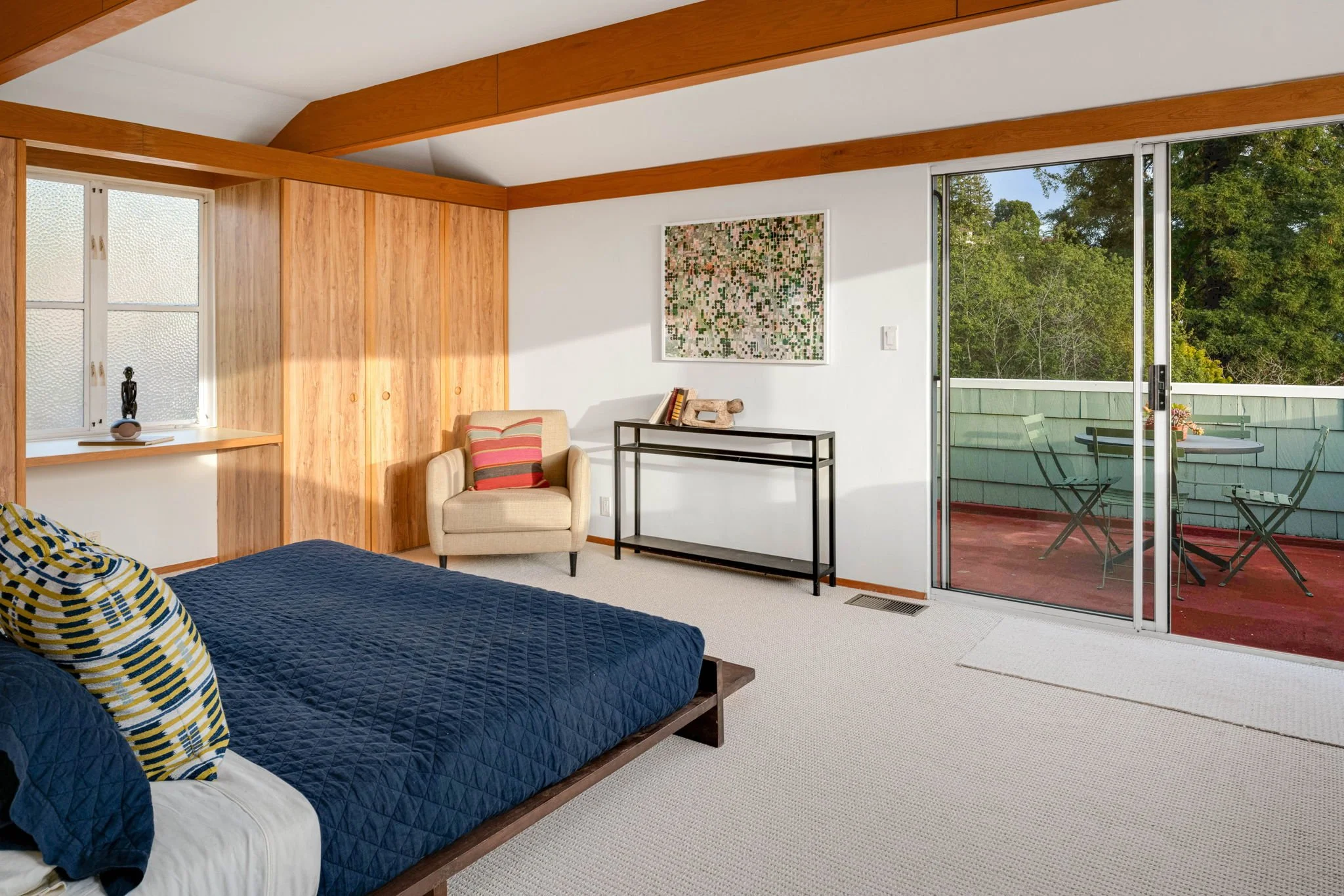 Bedroom with a bed, armchair, sliding glass door leading to a balcony with outdoor furniture, and window with a sculpture.