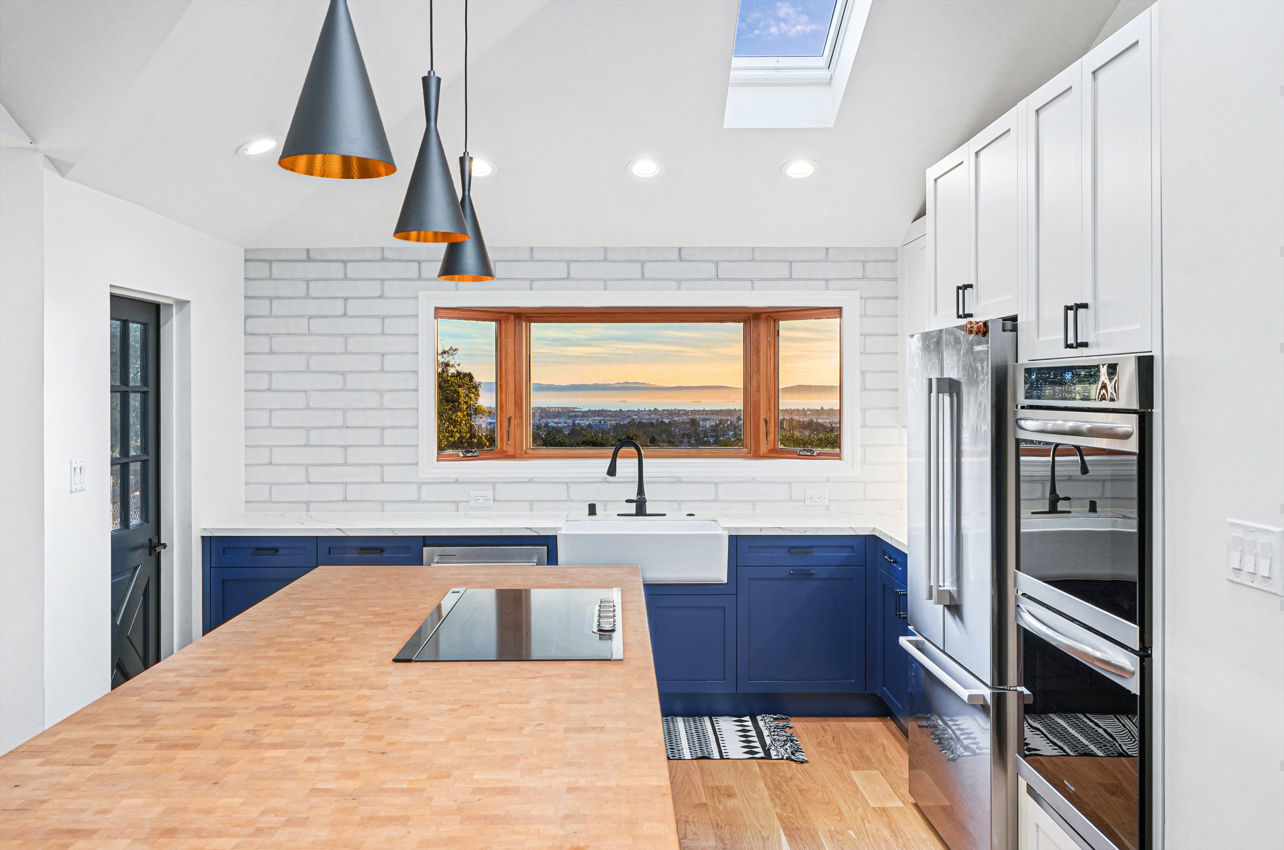 Modern kitchen with white brick walls, blue cabinets, stainless steel appliances, a large farmhouse sink, and a wooden countertop island. Ceiling skylight and a window offer a view of the sunset landscape.
