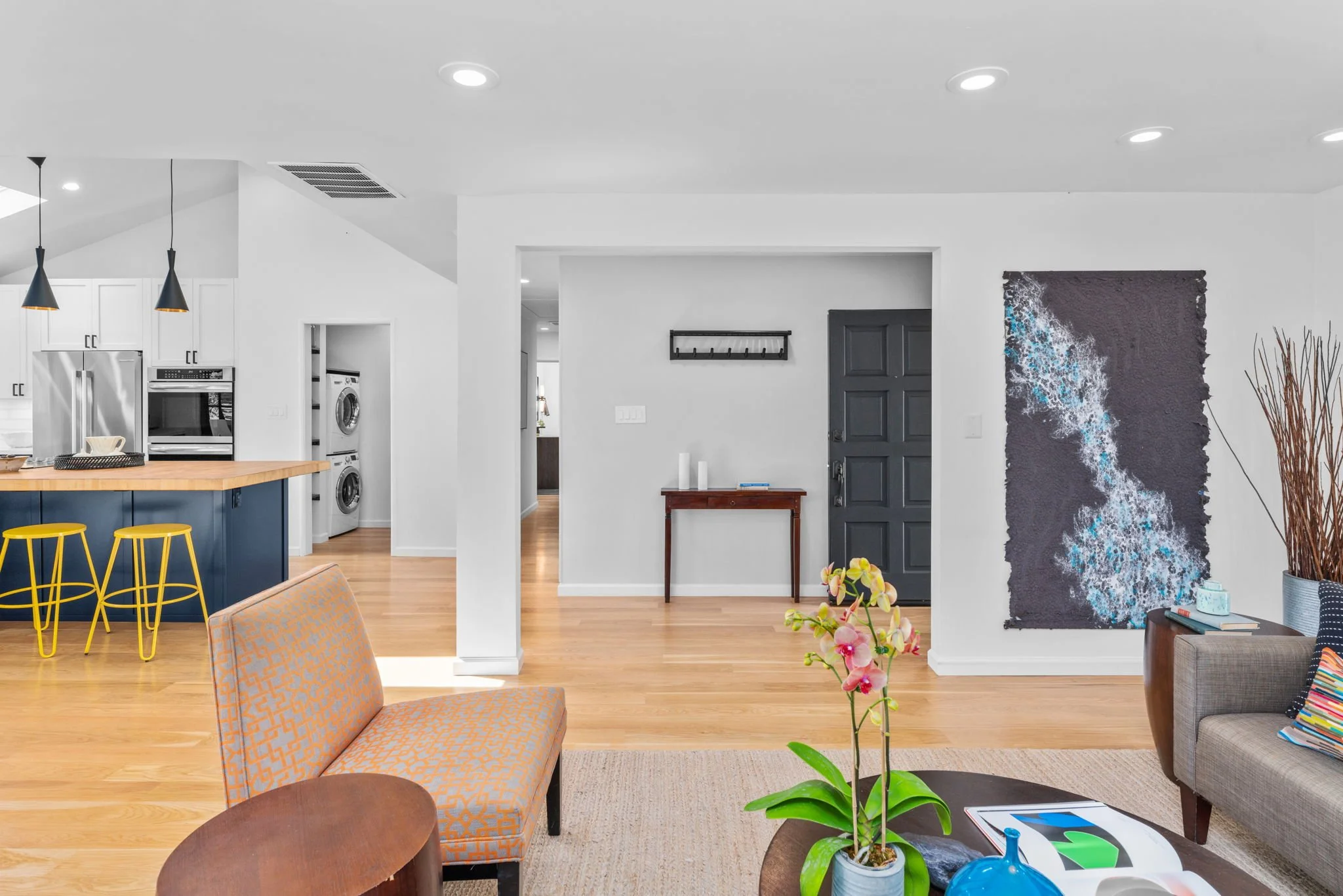 Living room with beige sofa, colorful throw pillows, coffee table with magazine and blue vase, potted orchid, abstract wall art, and modern decor. Open kitchen in background with black and yellow bar stools, white cabinets, and stainless steel appliances.