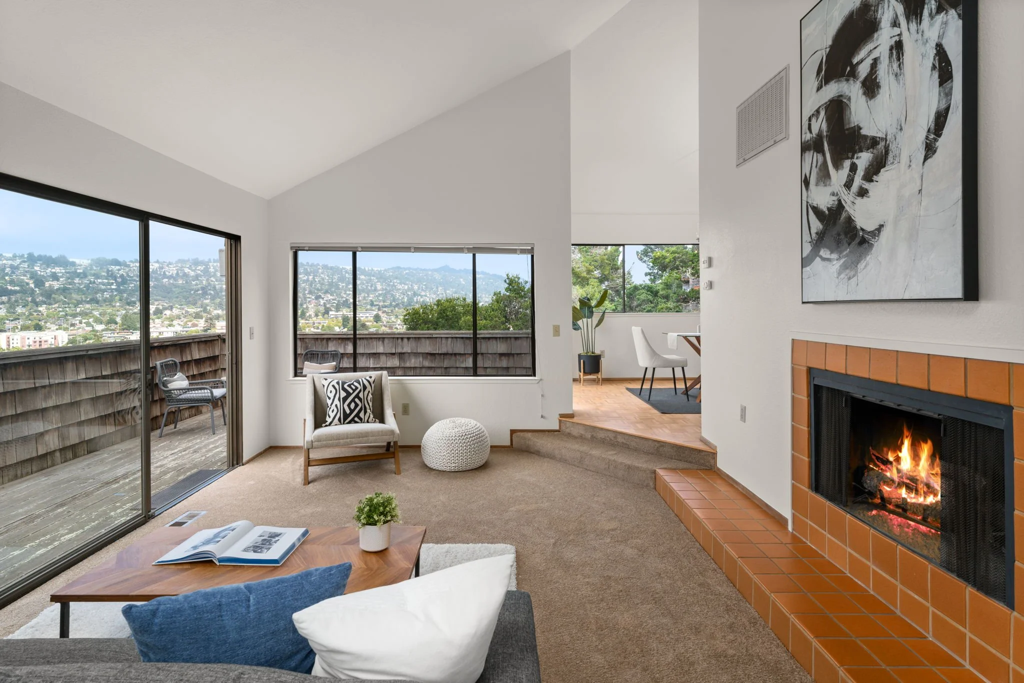 Living room with a fireplace, sofa, armchair, coffee table, and sliding glass door leading to a balcony with outdoor chairs. Large windows provide a view of a cityscape and hillside. There is a dining area in the background.