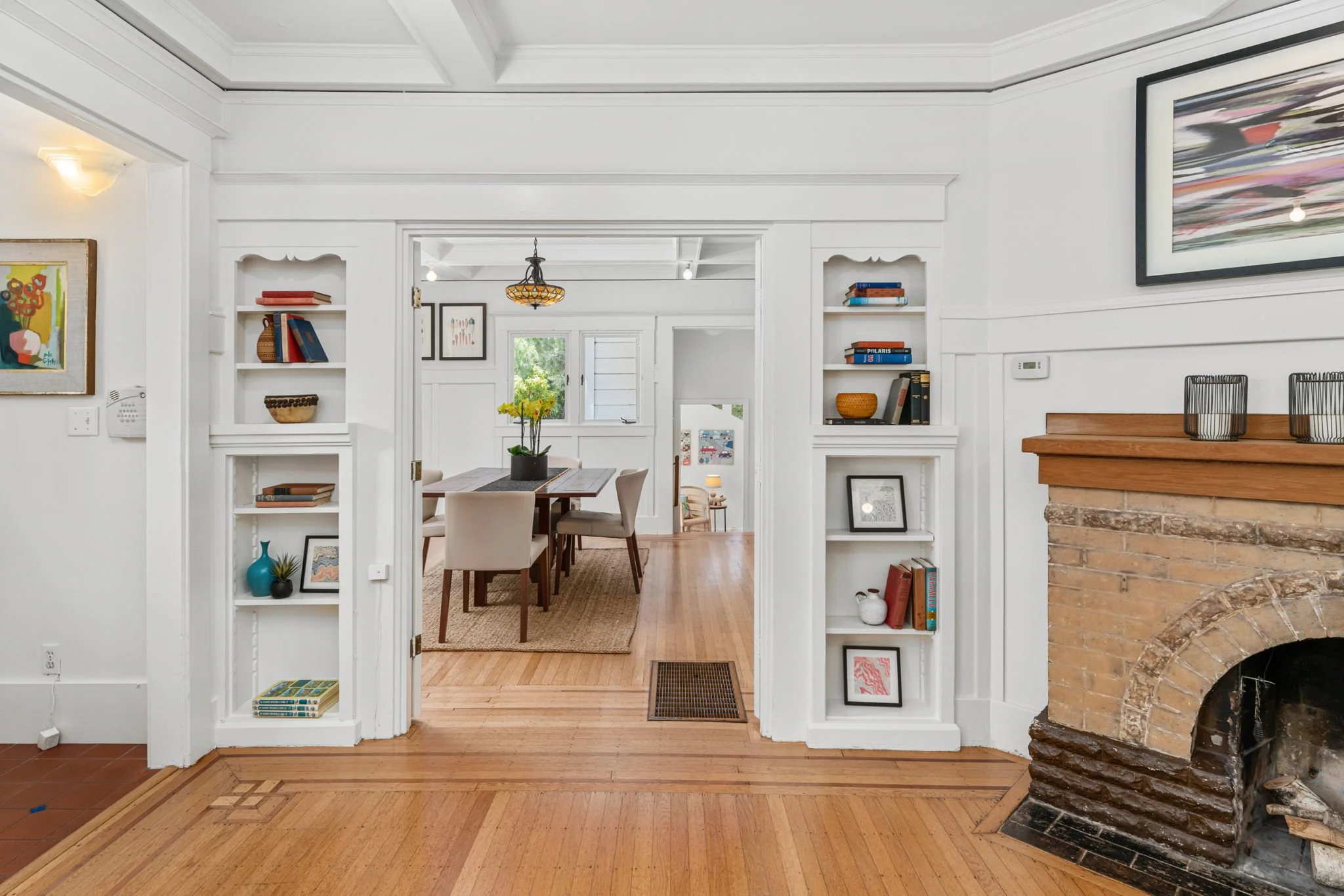 View of a bright, white living room leading into a dining area with wooden flooring, built-in bookshelves, and a brick fireplace on the right.