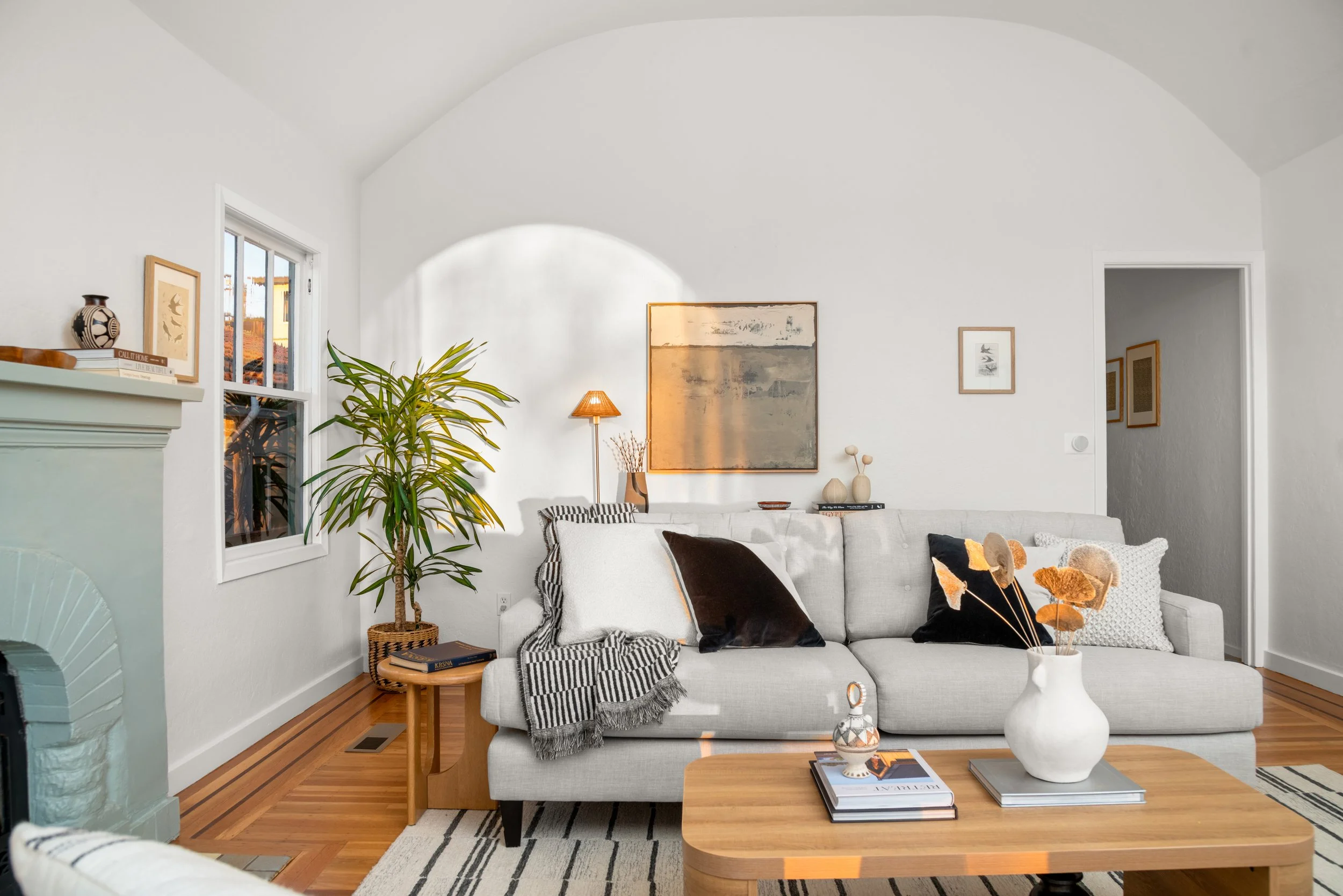 Living room with white walls, hardwood floors, a white sectional sofa with pillows, a coffee table with a vase and books, artwork on the wall, a tall lamp, a large plant near windows, and decorative items on the mantel.