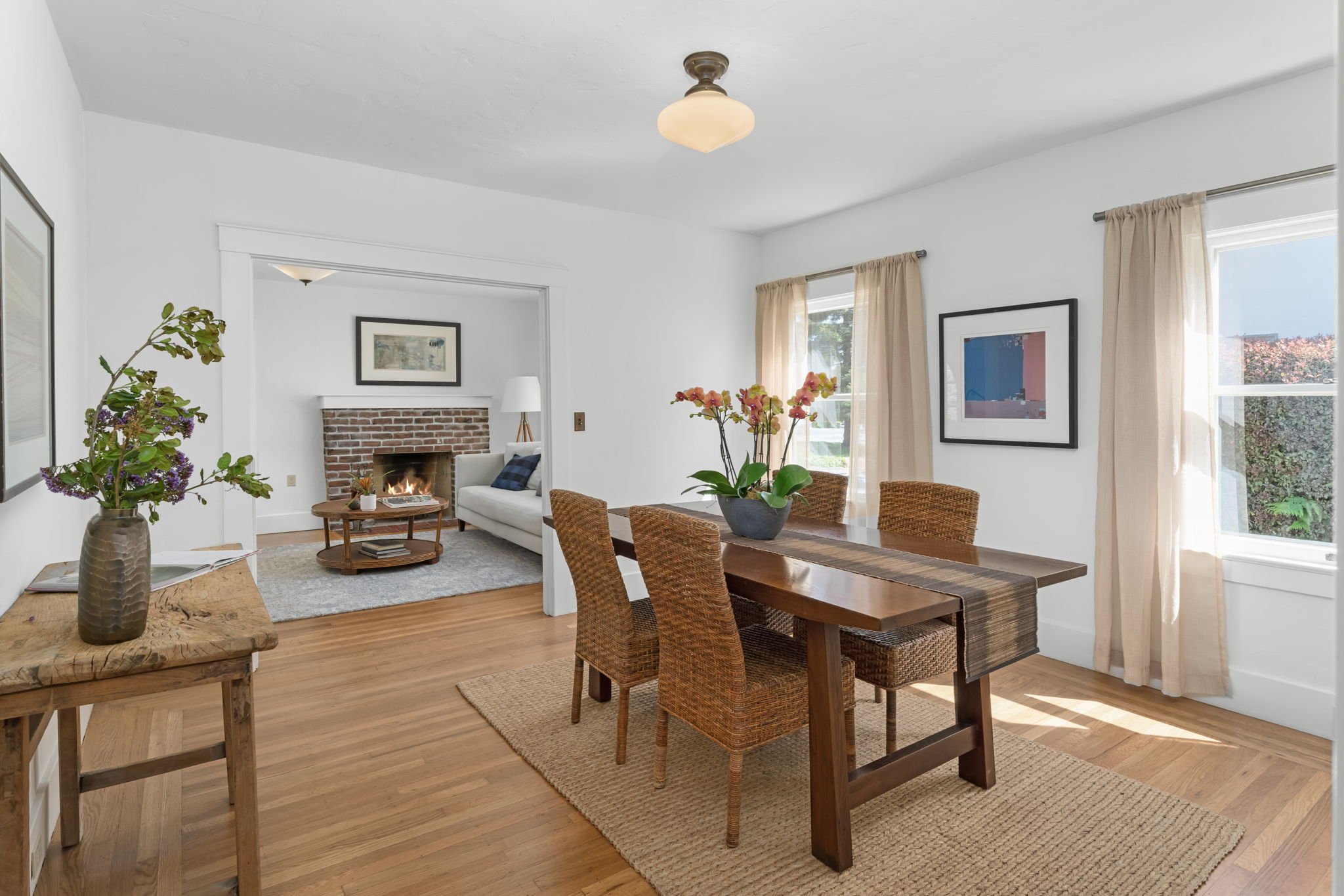 Dining room with wooden table, four chairs, pink orchids in a vase, and window with curtains. Connected to a living room with a brick fireplace, beige sofa, and framed art on the wall.
