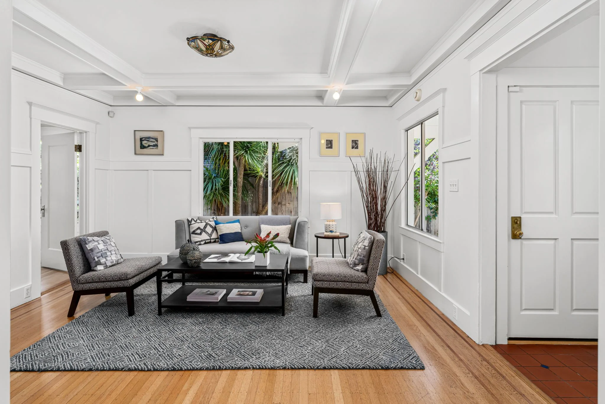 Living room with white walls, hardwood floors, a gray rug, and modern furniture including a gray sofa, patterned chairs, a black coffee table, and a side table with a lamp; large windows with greenery outside.