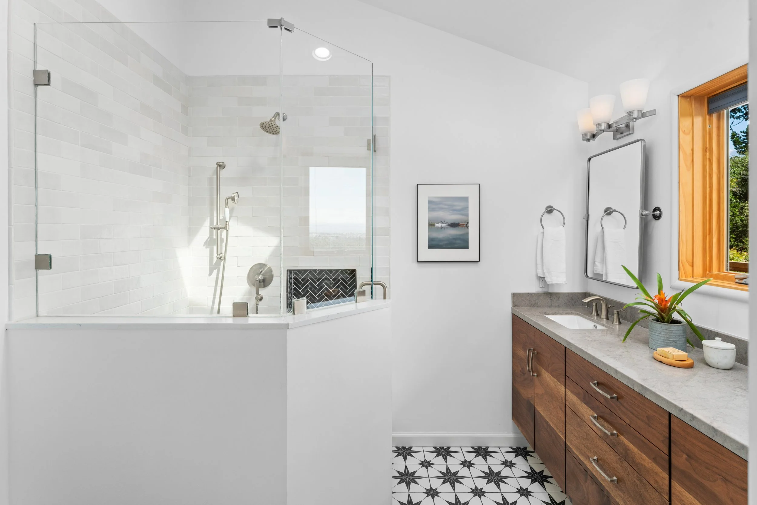 Modern bathroom with a glass-enclosed shower, white subway tiles, a wooden vanity with a gray countertop, a mirror, and a window showing greenery outside.