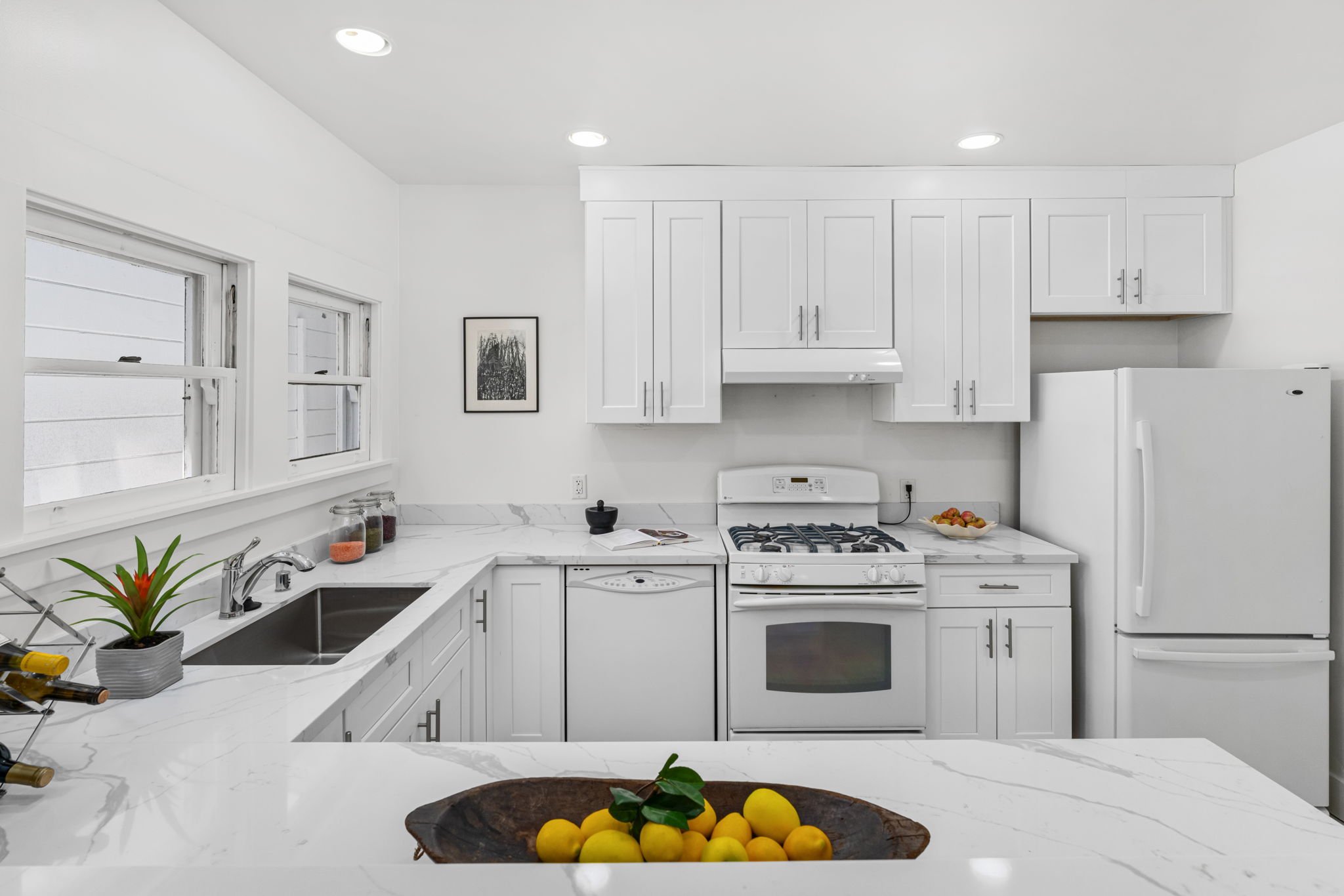 White kitchen with marble countertops, white cabinets, a refrigerator, stove, and microwave. Two windows above the sink, a black and white picture on the wall, a plant, and a bowl of lemons.