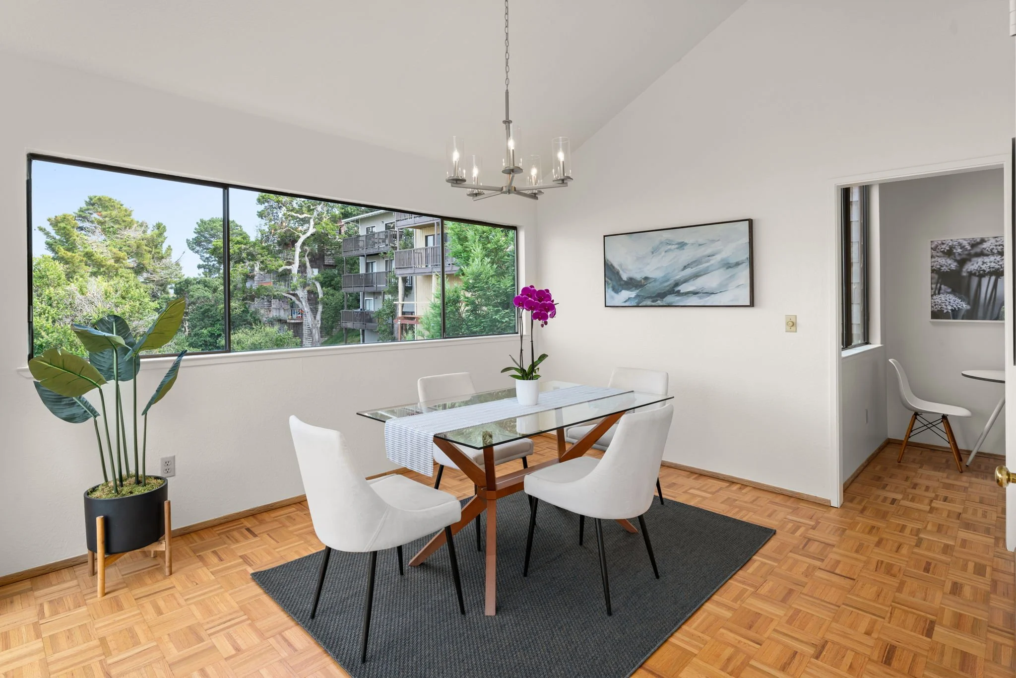 Dining room with white walls, wooden parquet floor, large window view of trees and buildings, glass-top table with a white table runner, five white chairs, a purple orchid in a white pot on the table, a black rug under the table, a modern chandelier,