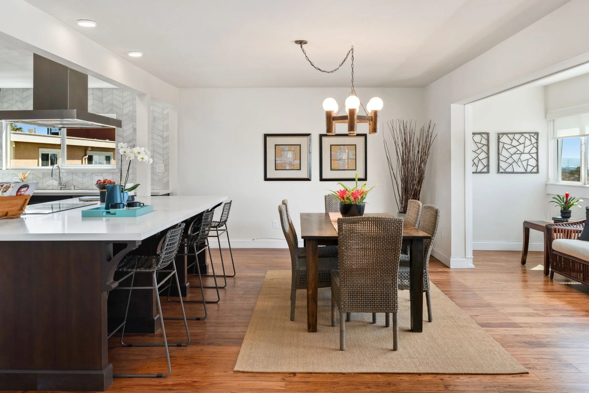 Open-concept dining and kitchen area with wooden floors, white walls, and bright natural light. Contains a kitchen island with bar stools, a wooden dining table with six chairs, decorative plants, and wall art.