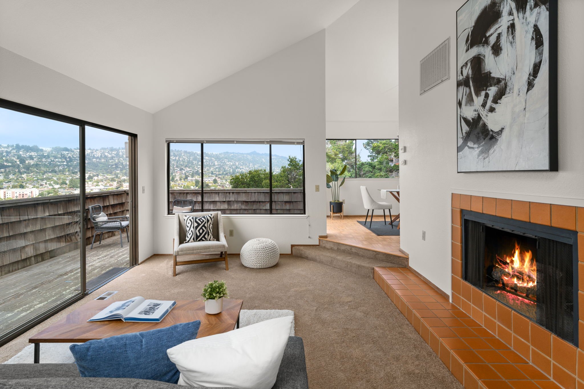 Living room with a brick fireplace, large windows with a view, a sofa with pillows, a coffee table with a book and plant, outdoor balcony with chairs, and a dining area in the background.