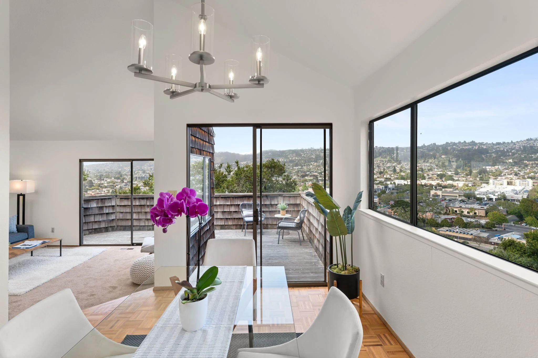 Interior of a modern apartment with large windows and a balcony, featuring a dining table with orchids, a living area with a lamp and sofa, and a view of a cityscape and hills outside.