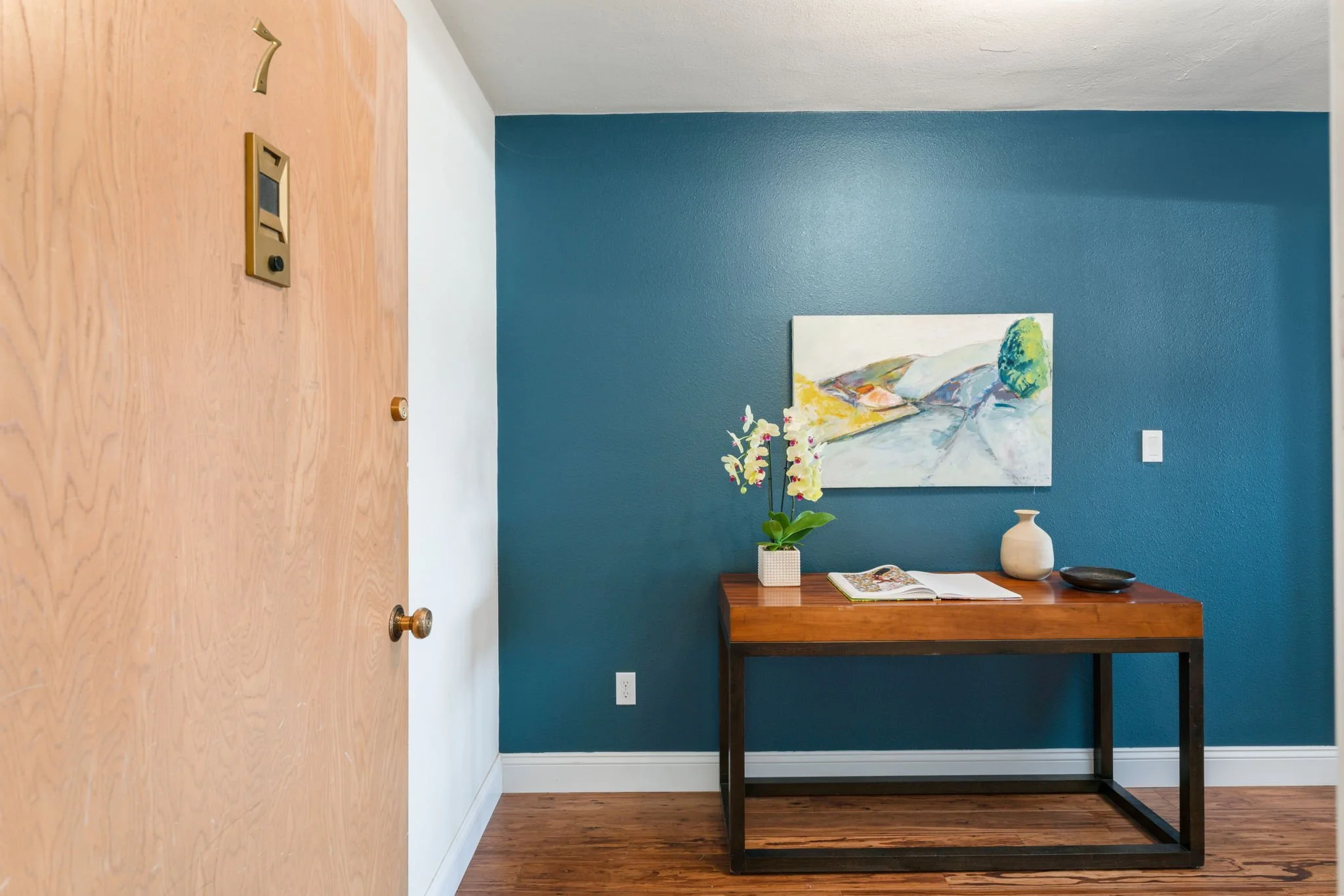 Interior of a room with a blue accent wall, wooden table with potted orchid, books, and ceramic vase, abstract landscape painting on the wall, and hardwood floor.