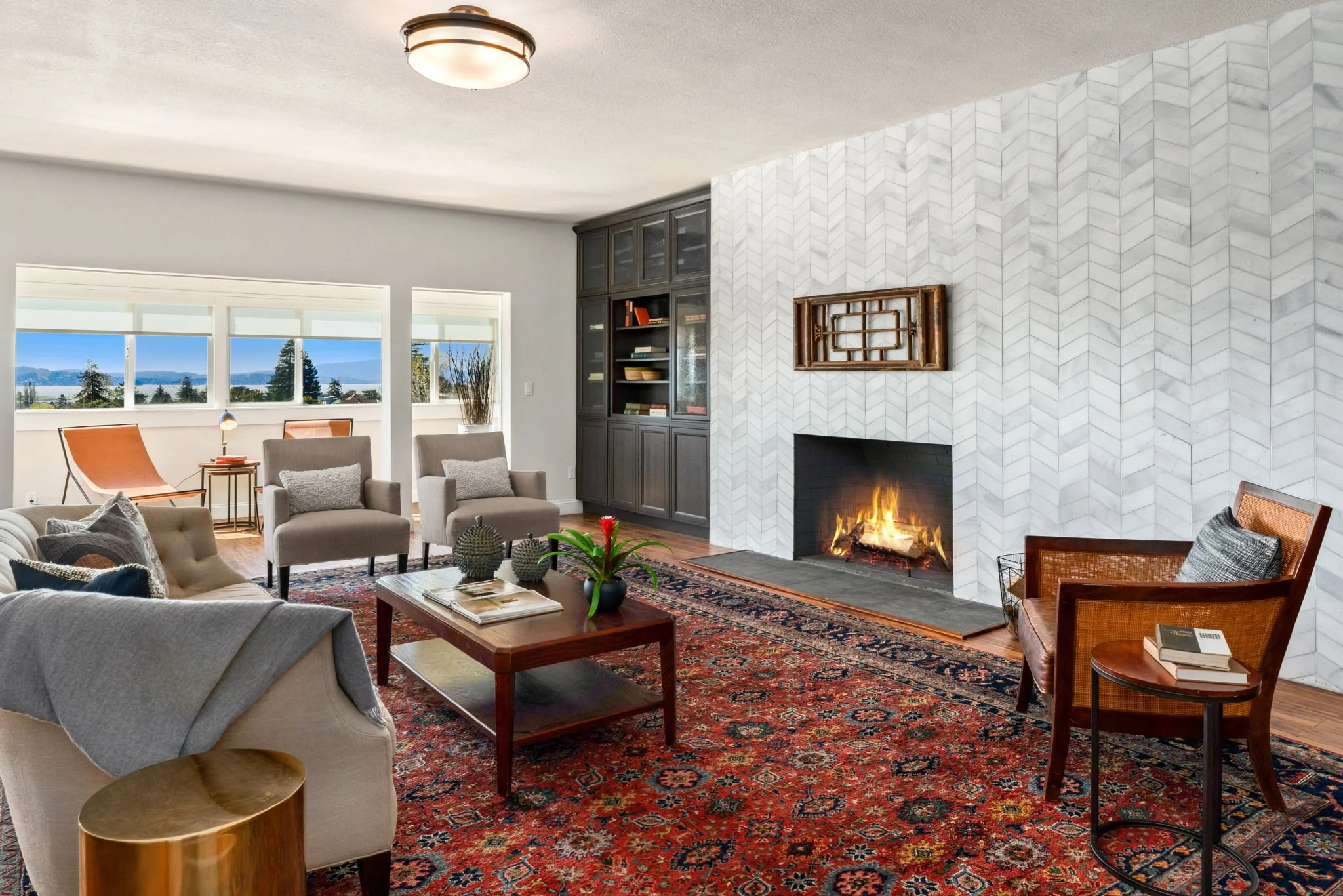 Living room with a fireplace, several chairs, a coffee table, and a large window with a view of trees and a body of water outside.