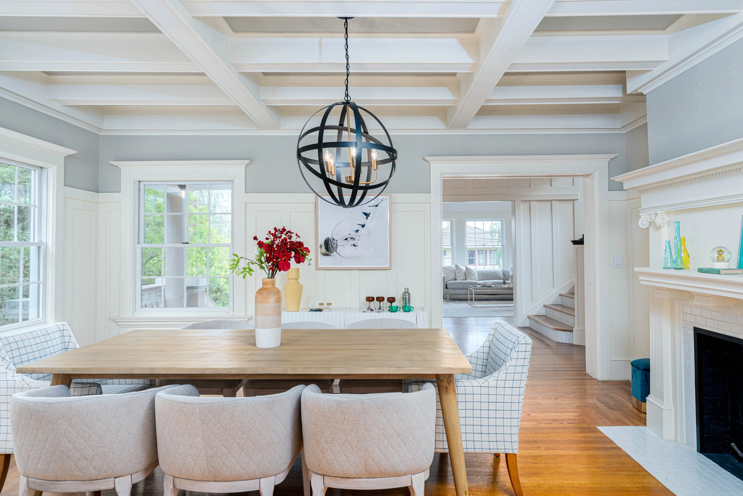 A spacious dining room with a wooden table surrounded by beige and checkered upholstered chairs, a large black spherical chandelier hanging from a white ceiling with exposed beams, and a fireplace with decorative glass bottles on the mantel. There ar