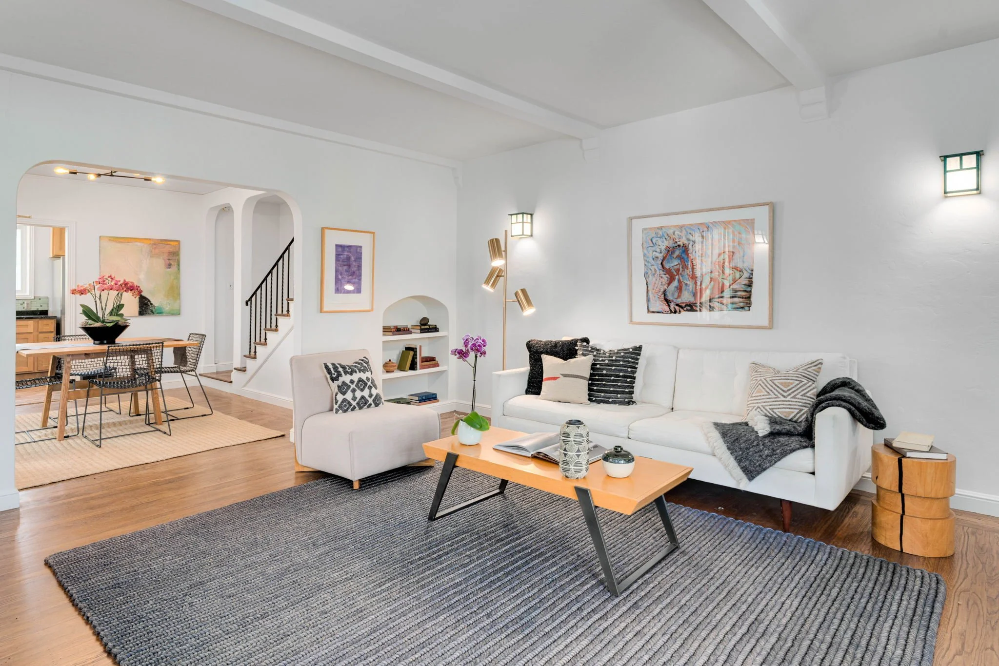 Living room with a white sofa, black and white pillows, a wooden coffee table with decorative items, a gray area rug, and artwork on the walls. Adjacent dining area with a table and chairs, and a staircase in the background.