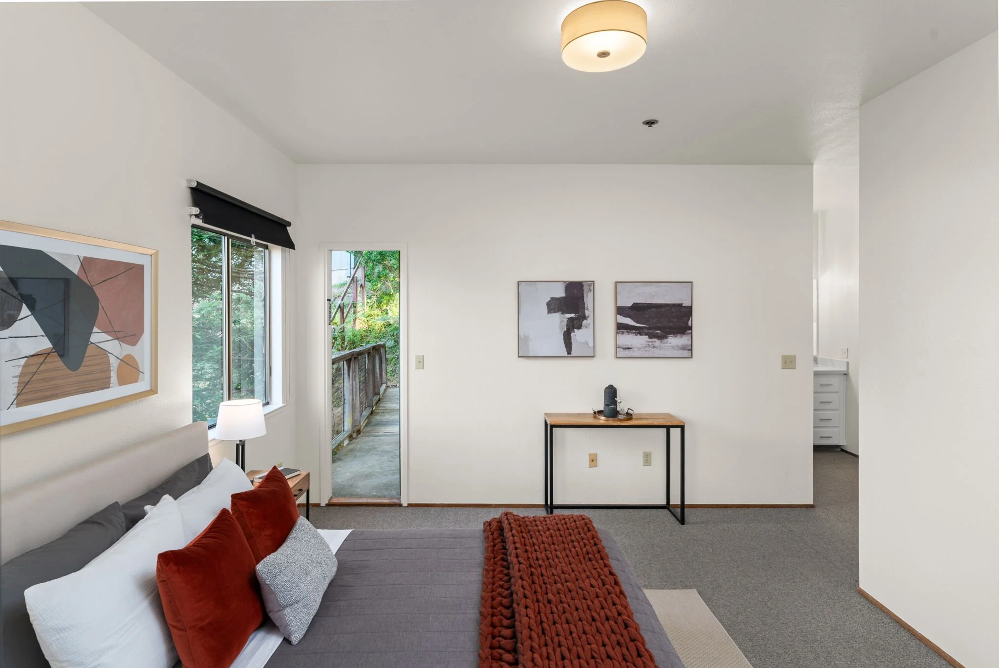 Interior view of a bedroom with a bed, pillows, a small side table with a lamp, decorated wall art, a small window, and a door leading to a balcony with greenery outside.