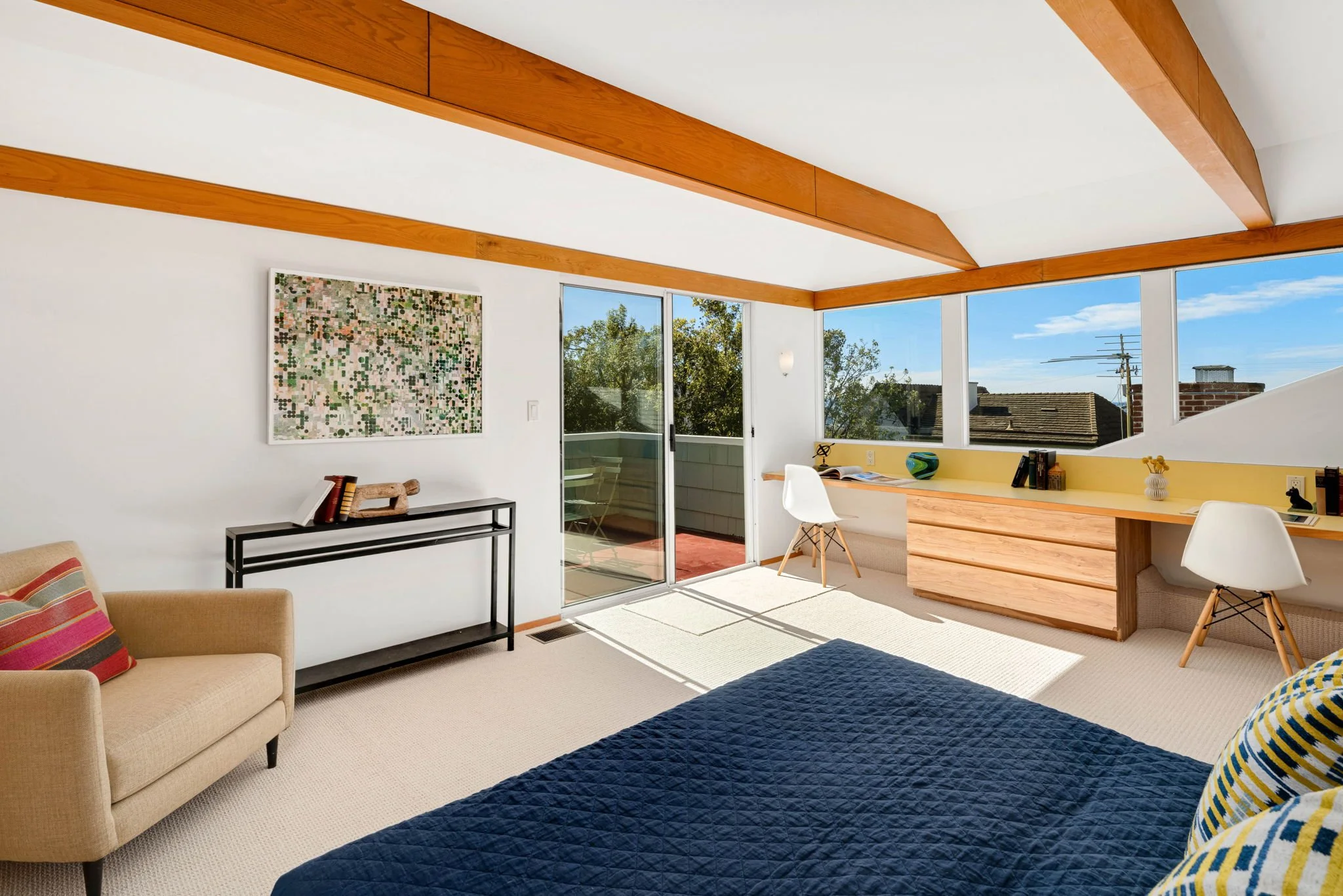 Bright bedroom with a beige couch, a black console table, abstract artwork, a sliding glass door opening to a small balcony, a built-in wooden desk with two white chairs, large windows showing a blue sky and trees, and a dark quilted bedspread.