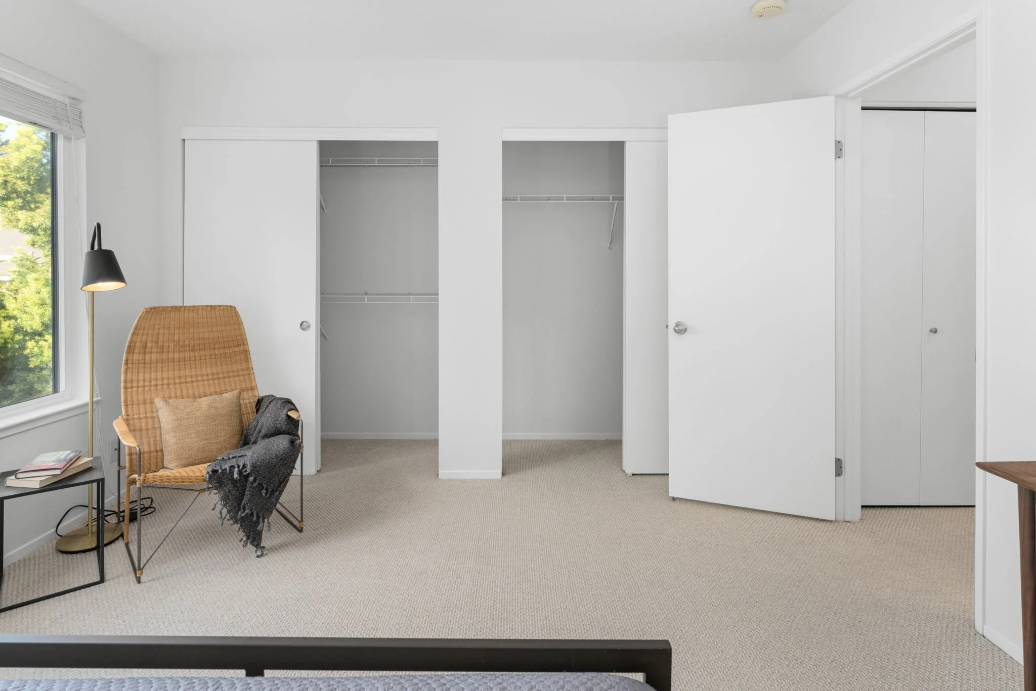 Empty bedroom with an open closet, a chair with a blanket and pillow, a side table with books, and a window with greenery outside.