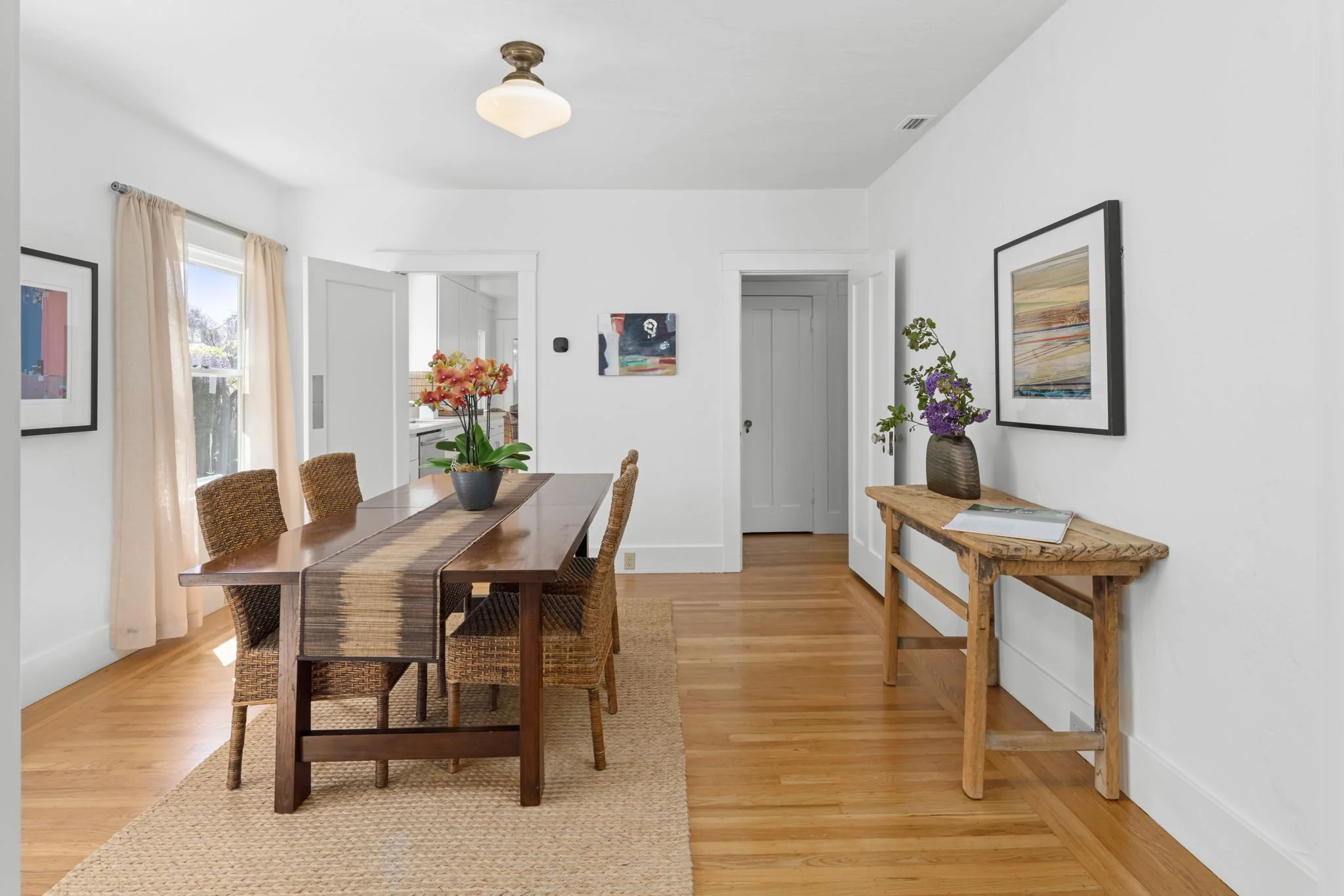 Dining room with a wooden table, six wicker chairs, a flower arrangement, wooden floor, and framed art on white walls.