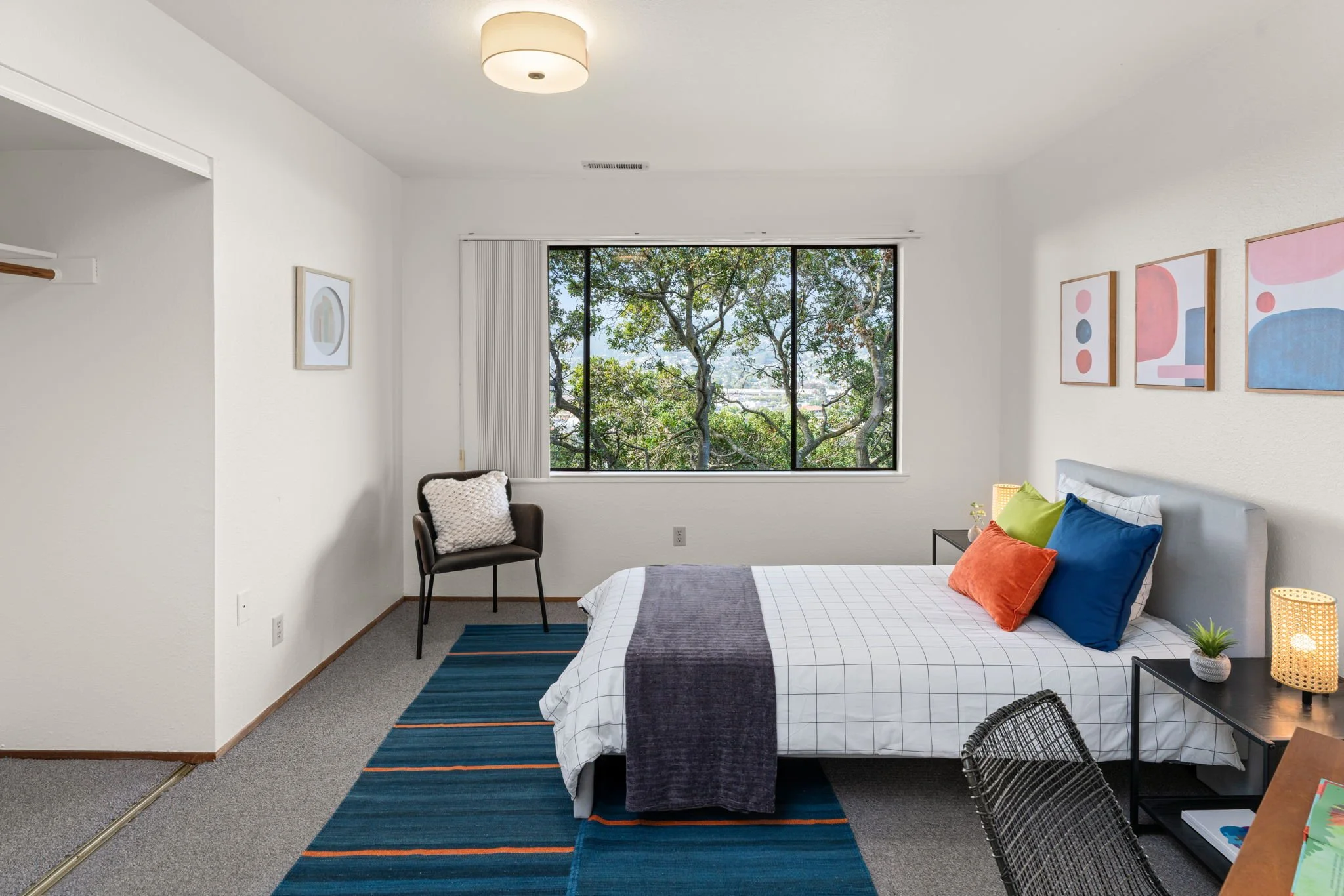 A bedroom with a large window showing trees outside, a bed with colorful pillows, a chair with a white pillow, a rug, and decorative wall art.