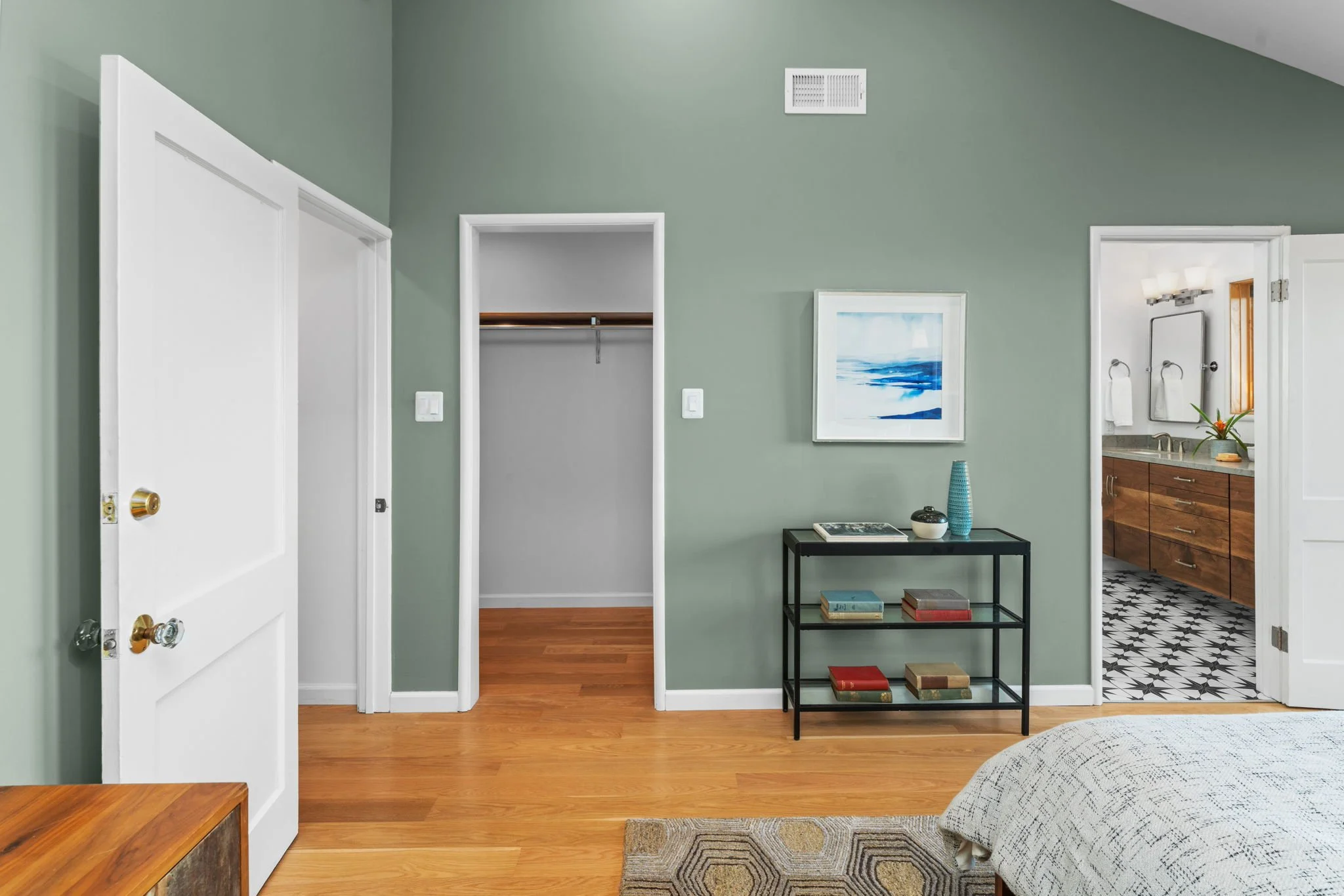 Bedroom with green wall, open closet, black console table with books and ornaments, artwork, doorway to bathroom with wooden vanity and patterned tiles.