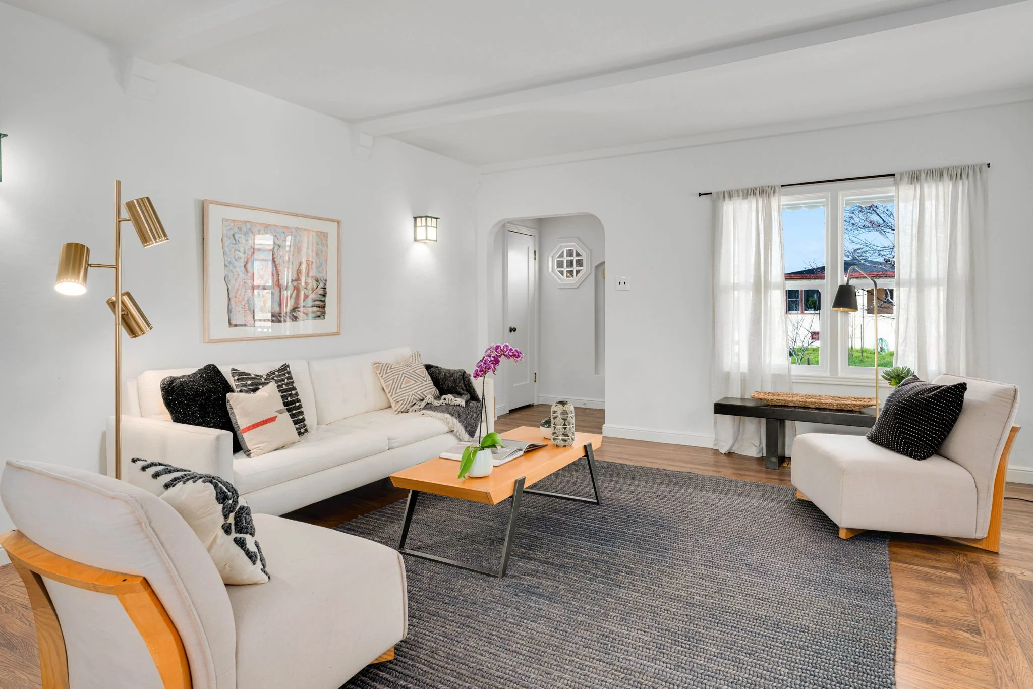 Bright living room with white sofa, armchair, black area rug, wooden coffee table with pink flowers, and large windows with white curtains.