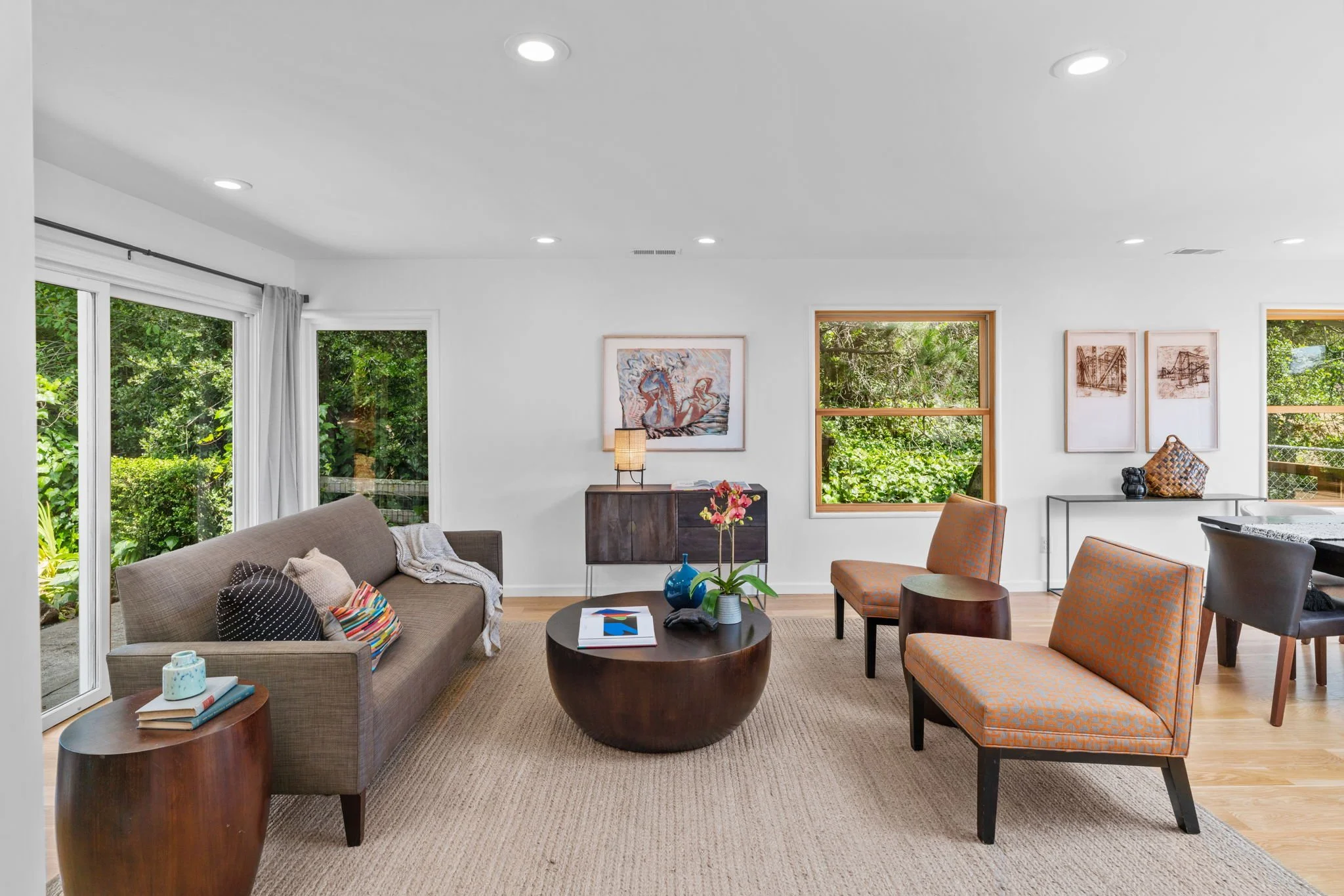 Living room with large windows, brown sofa with cushions, round wooden coffee table with flowers, two upholstered chairs, side table with books, framed artwork on white walls, and outdoor greenery visible through windows.