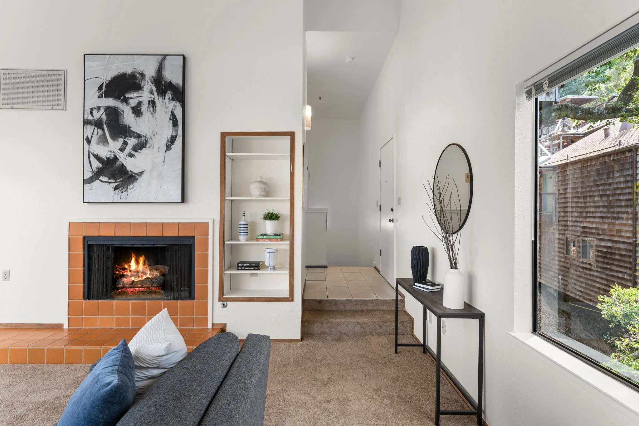 Living room with a gray sofa, white walls, a fireplace with orange tiles, a black-and-white abstract painting above the fireplace, a built-in white shelf with decorative items, a console table with a vase and books, a  large window showing an outdoor