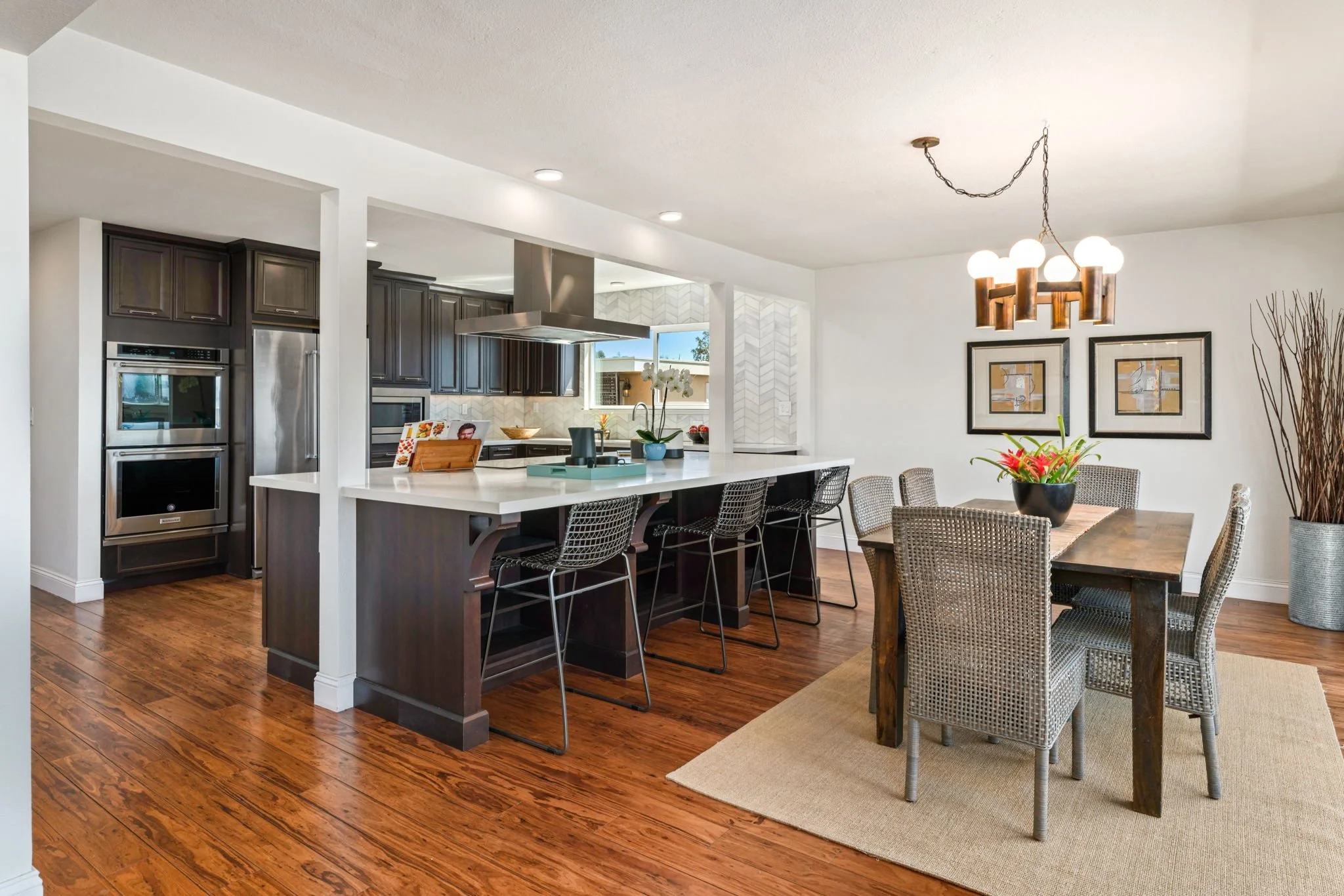 Open-concept kitchen and dining area with dark wood cabinetry, stainless steel appliances, white countertops, a breakfast bar with black wire chairs, a rustic dining table with wicker chairs, a chandelier, framed artwork, a large potted plant, and ha