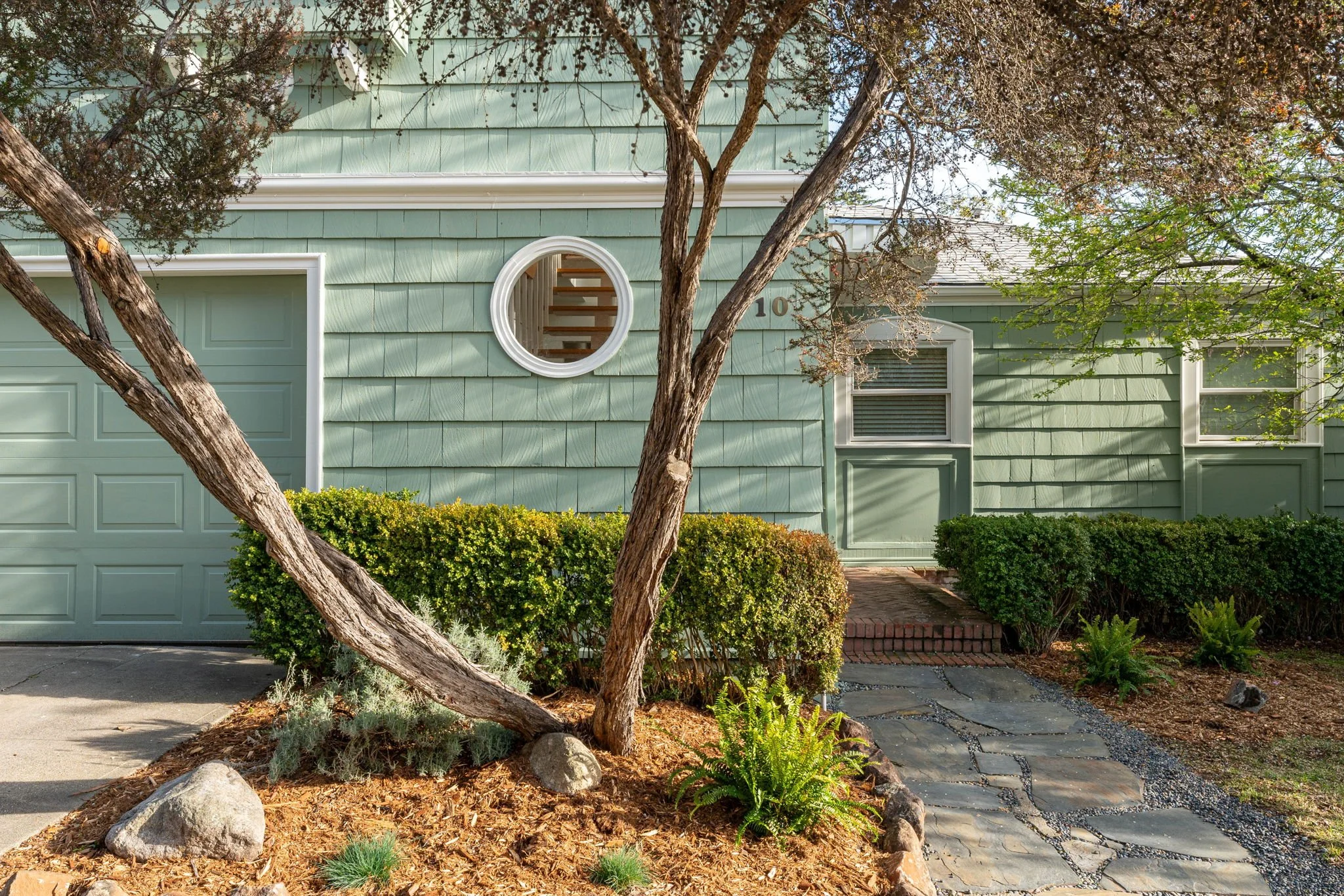 Front view of a light green house with a garage door, a round window, a rectangular window with shutters, brick steps, a pathway, trees, trimmed bushes, and landscaped garden.