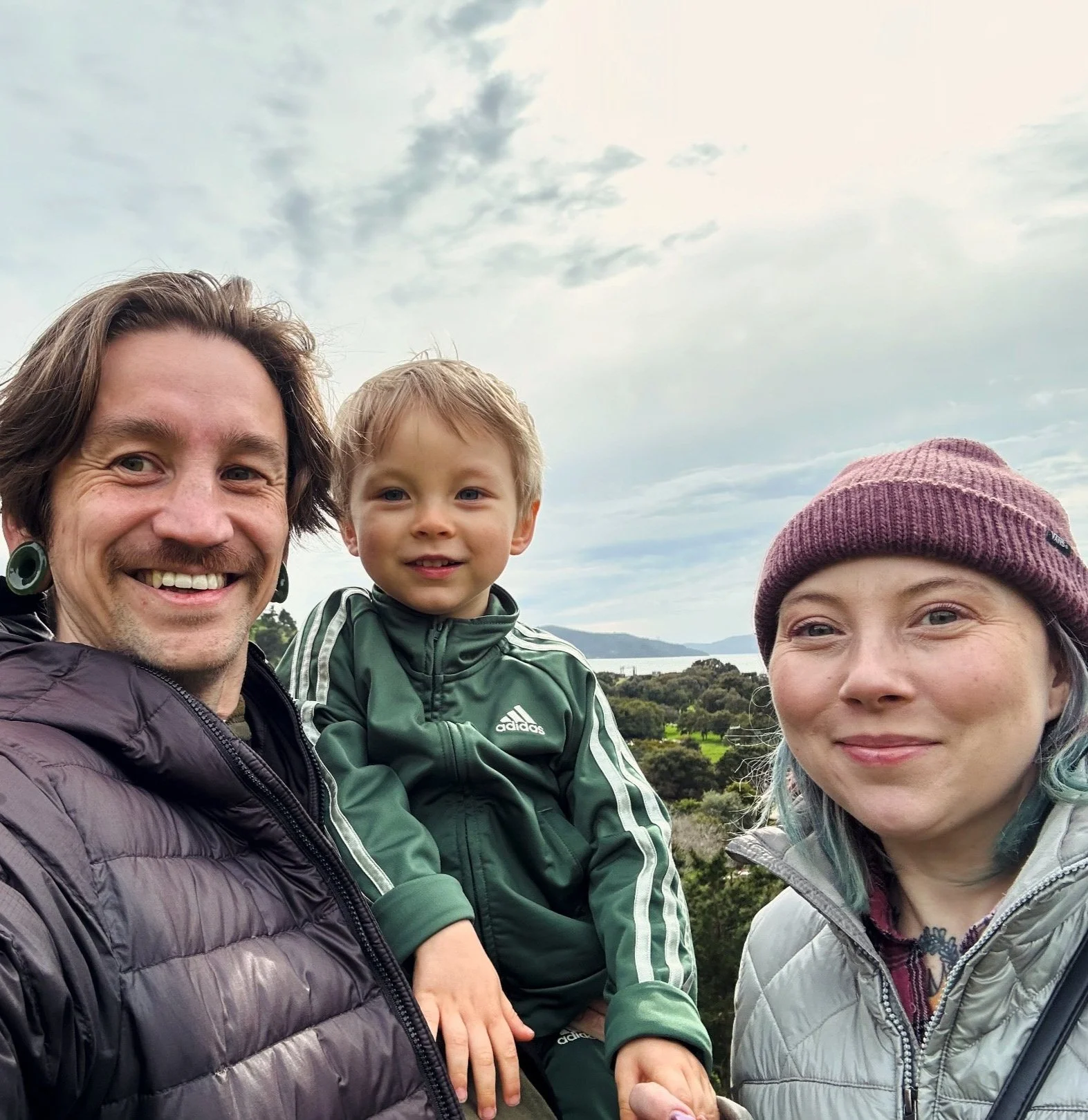 A family portrait outdoors with a scenic background of water and trees, featuring two adults and a young boy, all smiling.