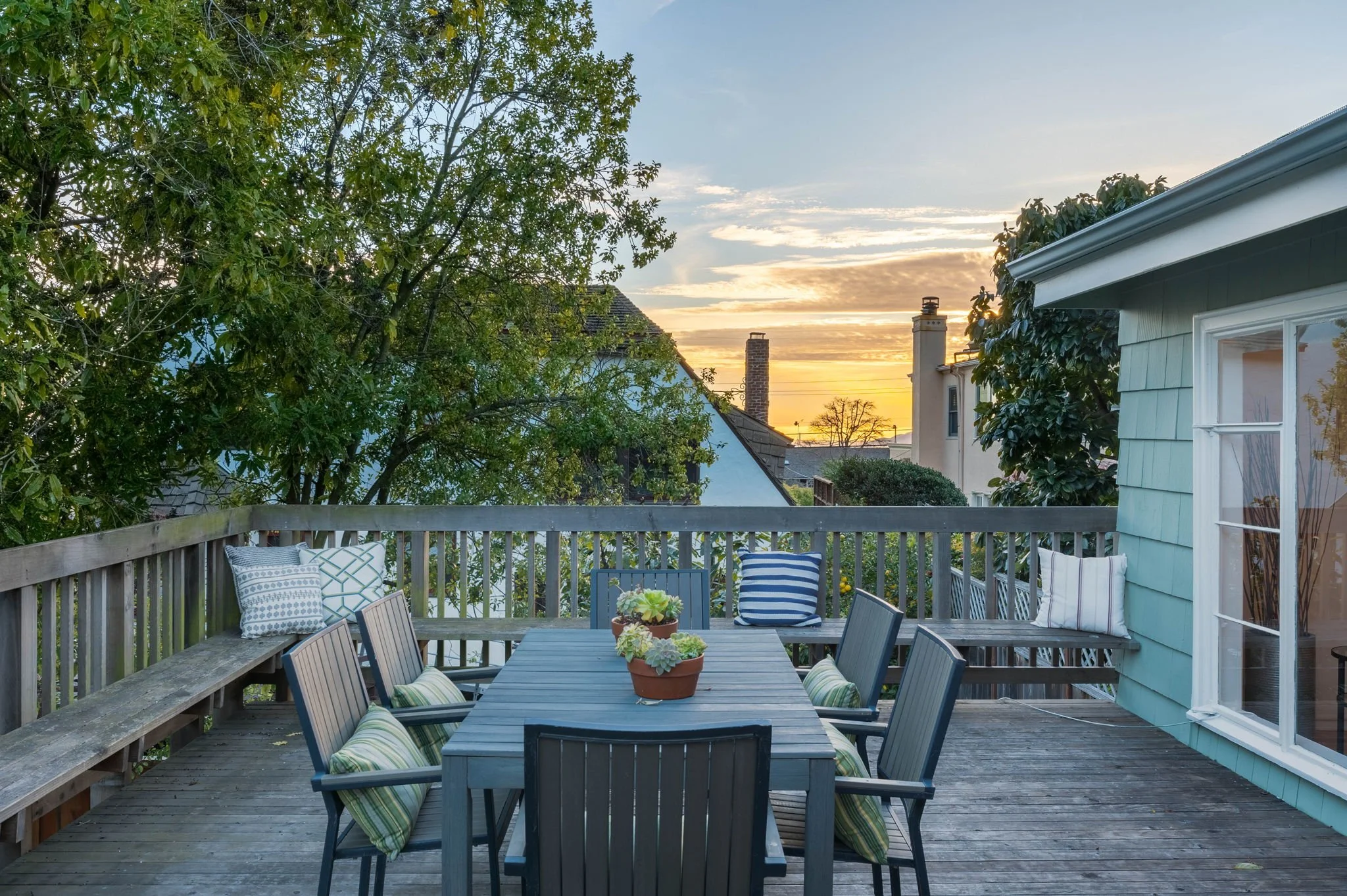 A wooden outdoor deck with six chairs around a rectangular table, decorated with striped and patterned cushions, surrounded by a wooden railing, with potted succulent plants on the table, and a sunset sky in the background.