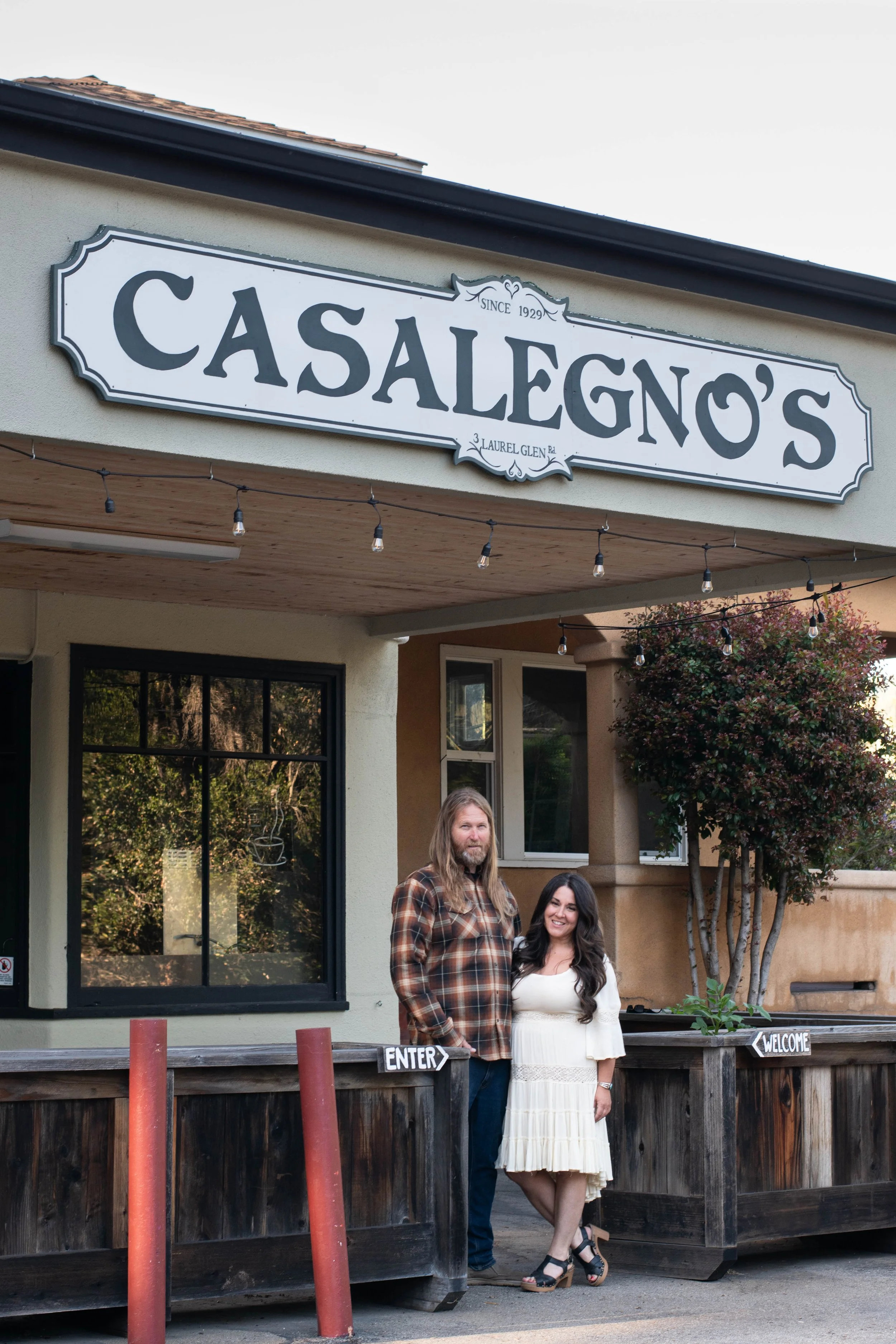 Two people standing in front of a restaurant with a sign that says "Casalegno's" and a wooden fence with signs that say "Enter" and "Welcome."