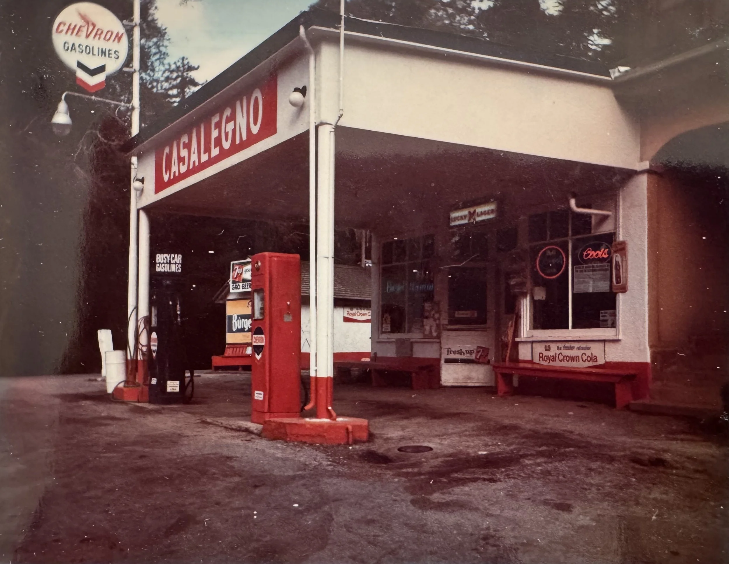An old vintage gas station with Chevron gasoline sign, a red gas pump, and a small snack shop called Casalegno. There is also a Royal Crown Cola sign visible behind the window.